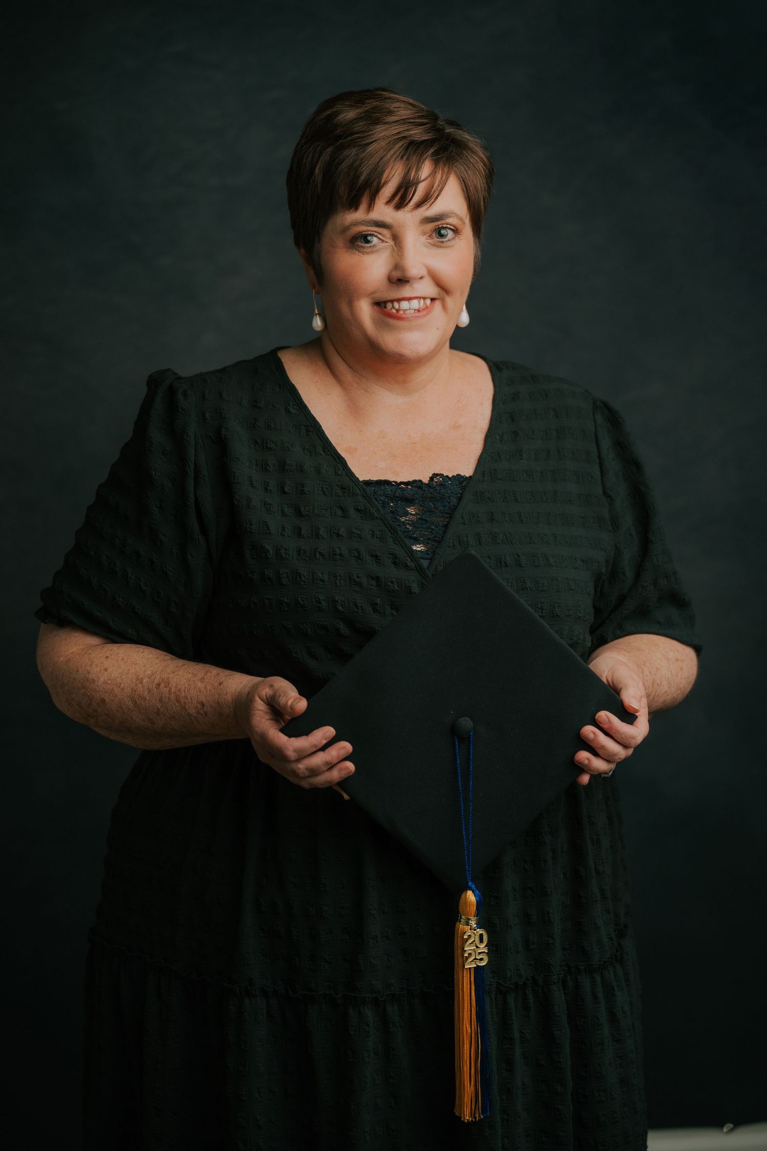 Graduate wearing black academic gown with gold tassel poses against dark background in professional portrait series.