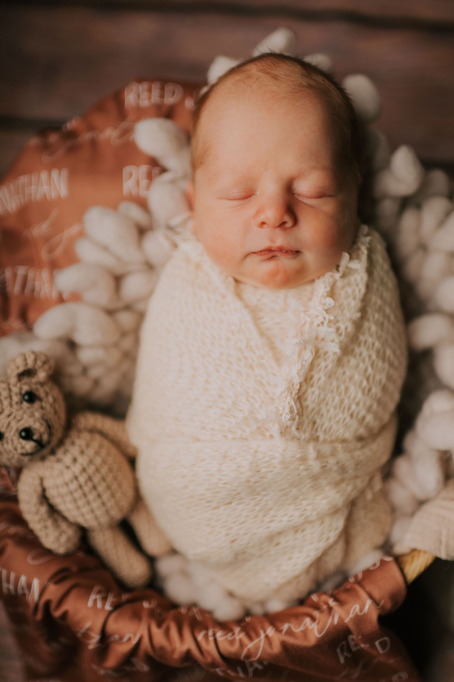 Newborn baby wrapped in white knit blanket with stuffed bear beside them.