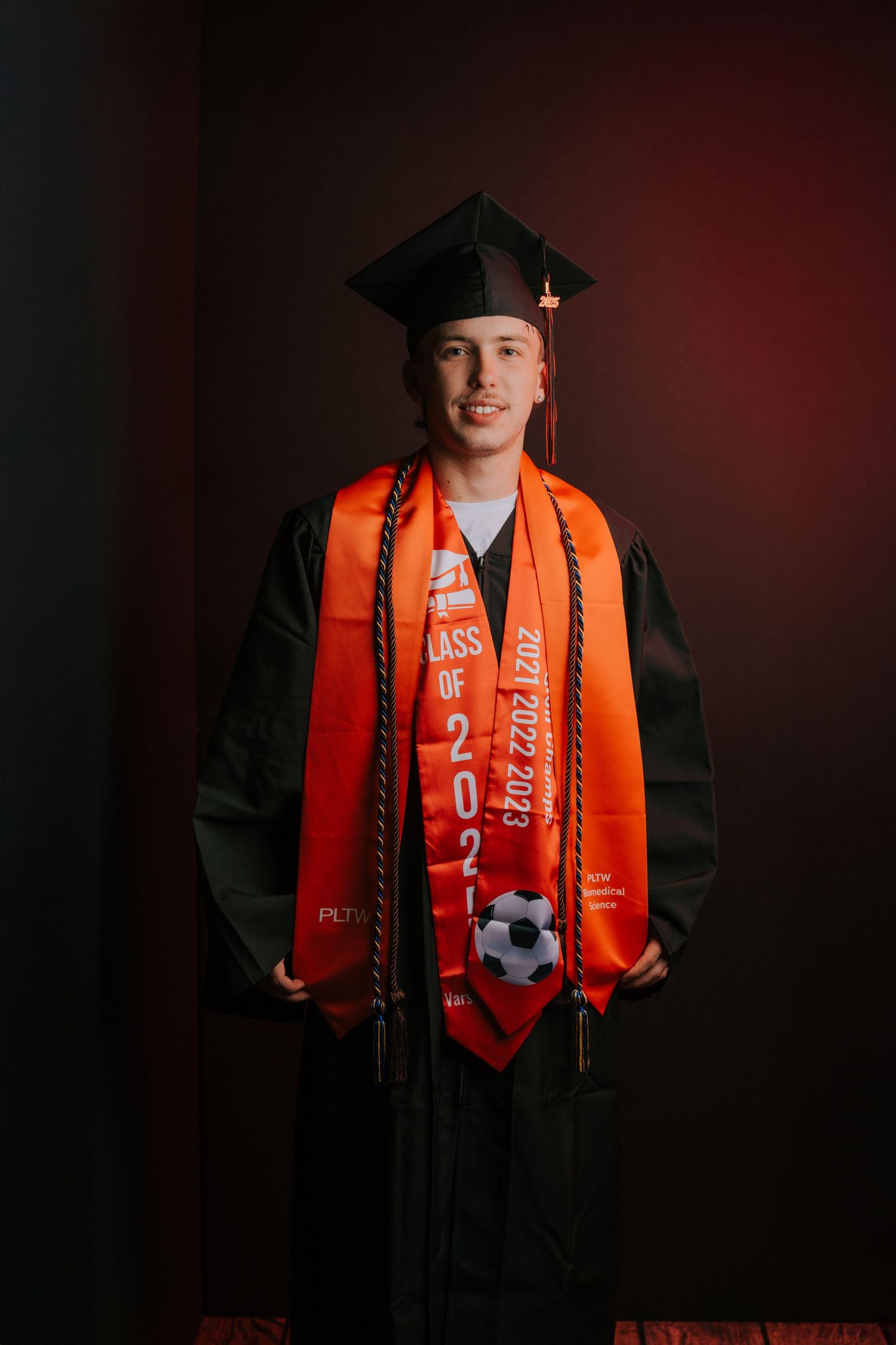 Graduate in cap and gown with orange stole poses against dark dramatic backdrop.