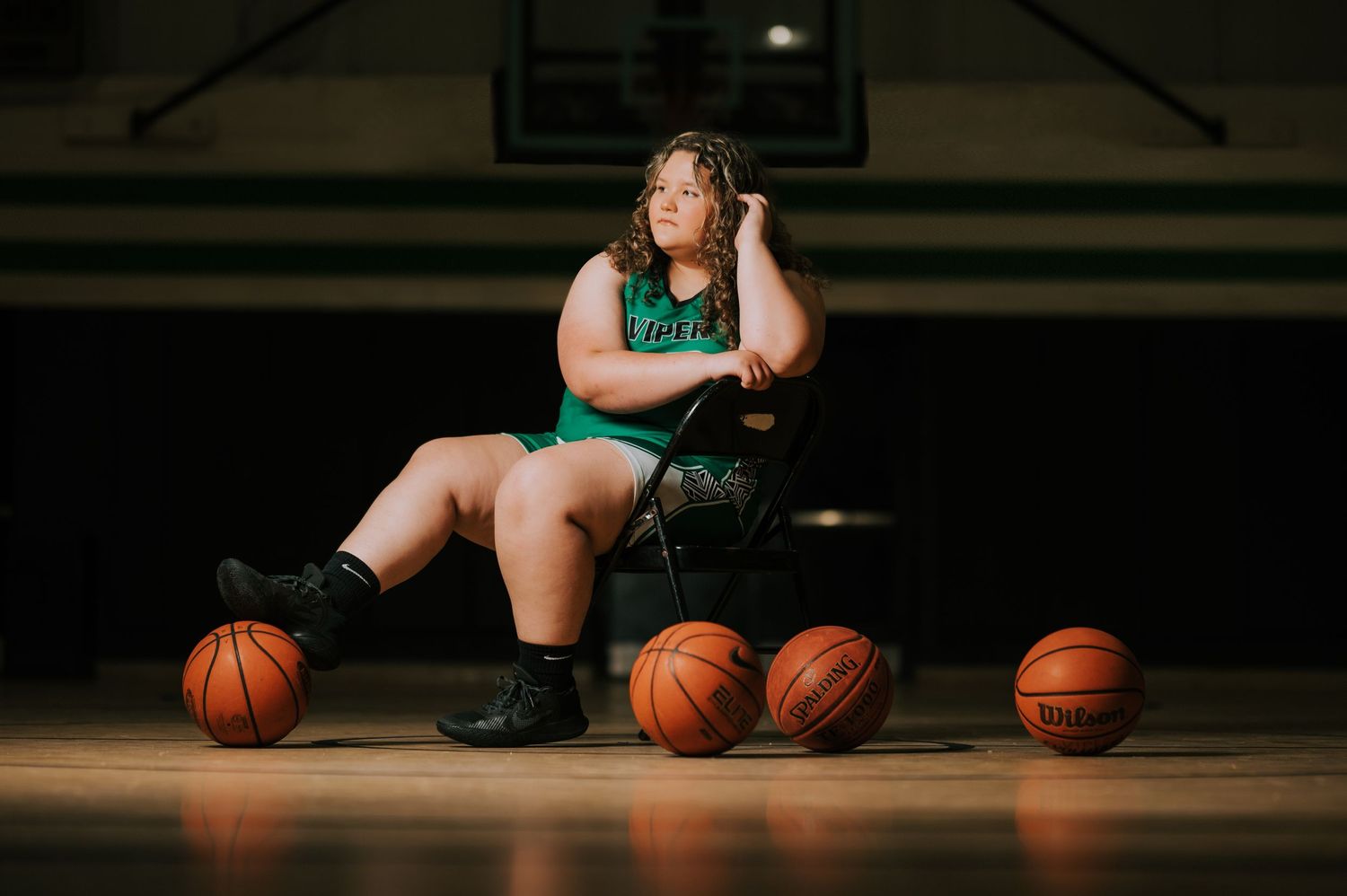 A basketball player sits on a wooden gym floor surrounded by multiple basketballs in dramatic lighting.