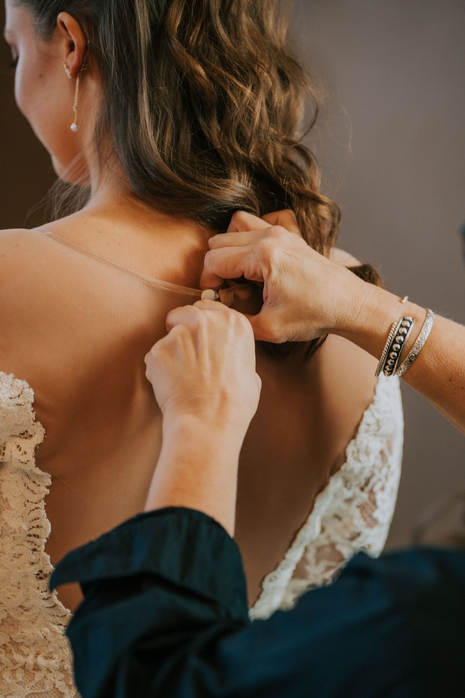Hands fastening the back of a delicate lace wedding dress with wavy brown hair visible.