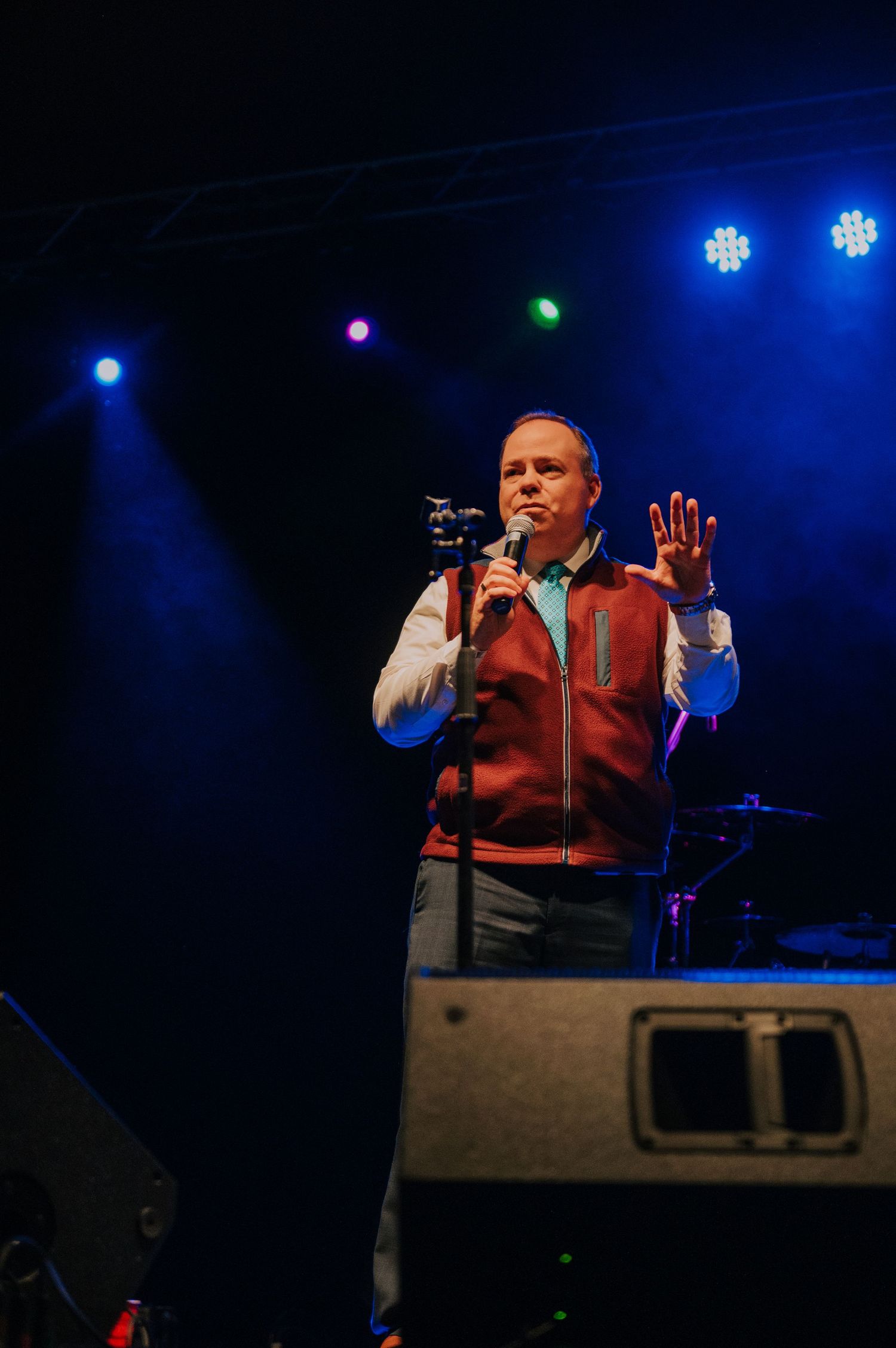 A performer wearing a red vest and grey pants sings on stage with colorful stage lighting in the background.