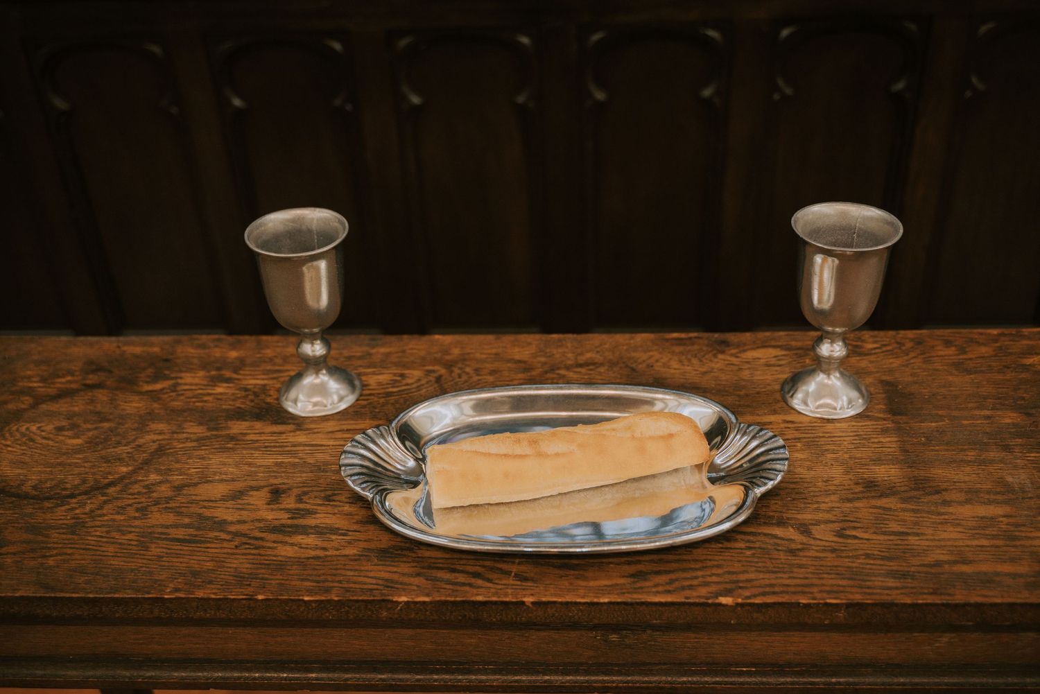 Two silver chalices and a slice of bread on a silver plate rest atop a rustic wooden table.