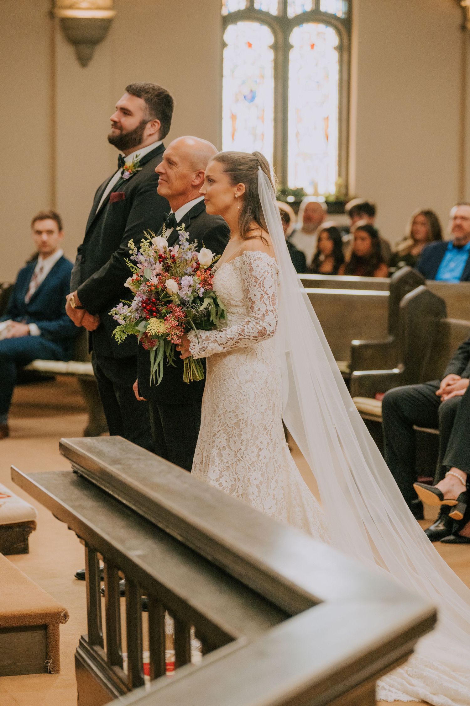 A traditional wedding ceremony in a church with stained glass windows as a couple stands at the altar with beautiful florals.