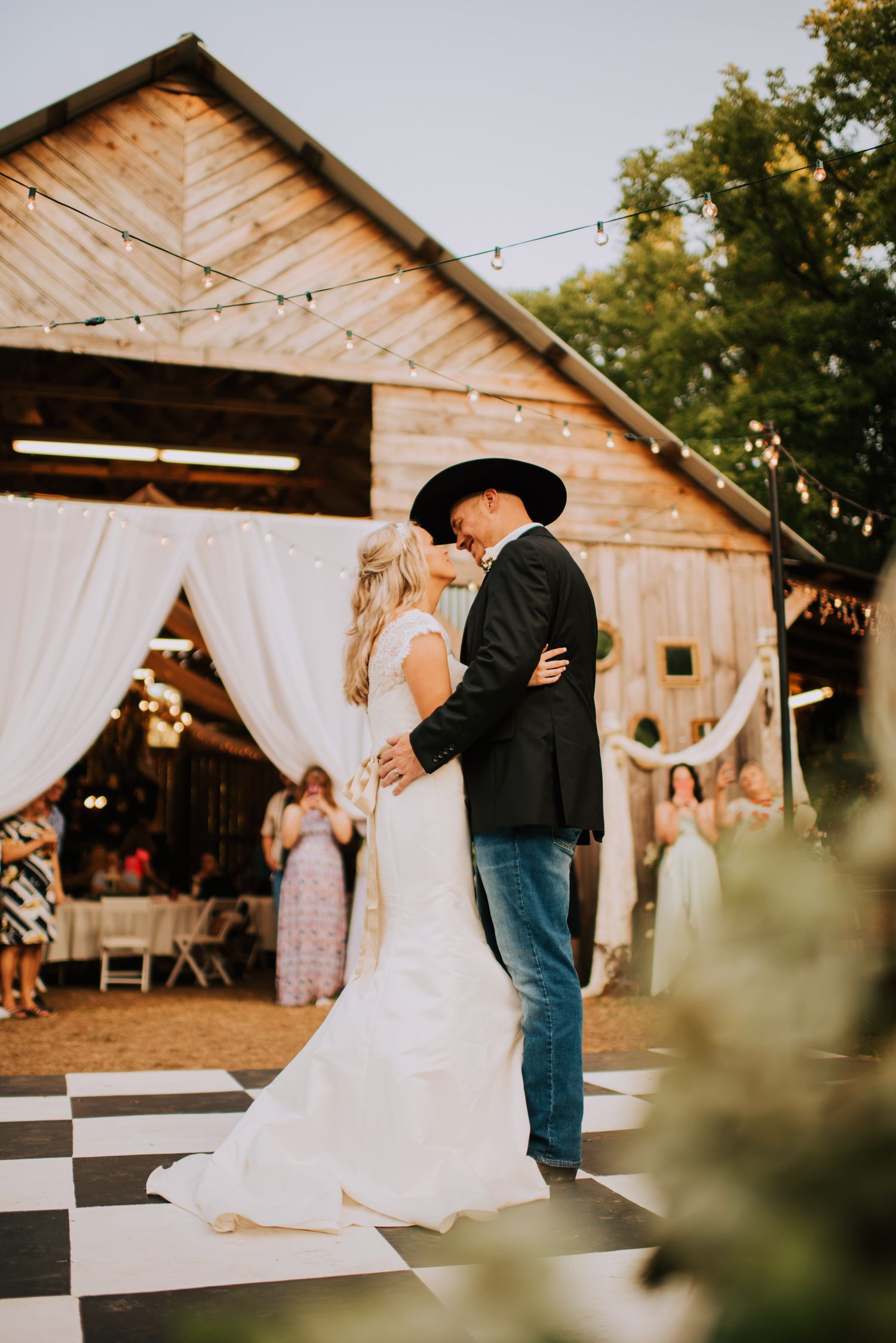A rustic barn wedding reception features couples dancing under string lights and white drapes.