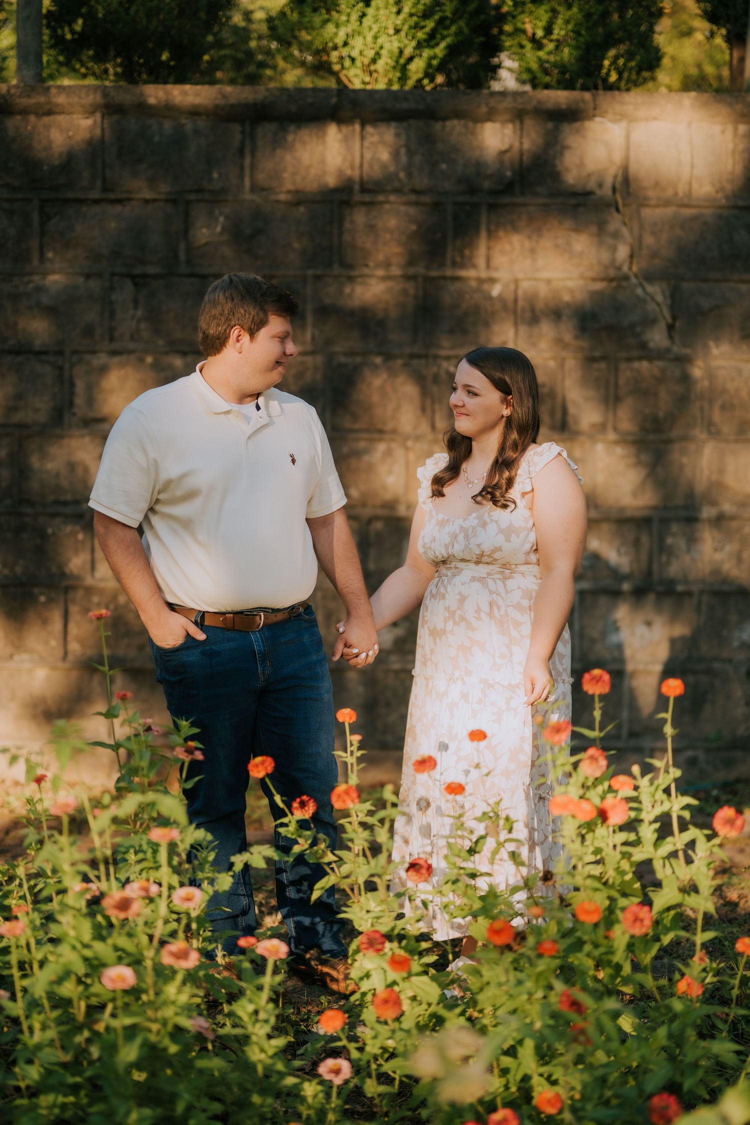 A couple walking together through a garden with blooming orange flowers against a stone wall.