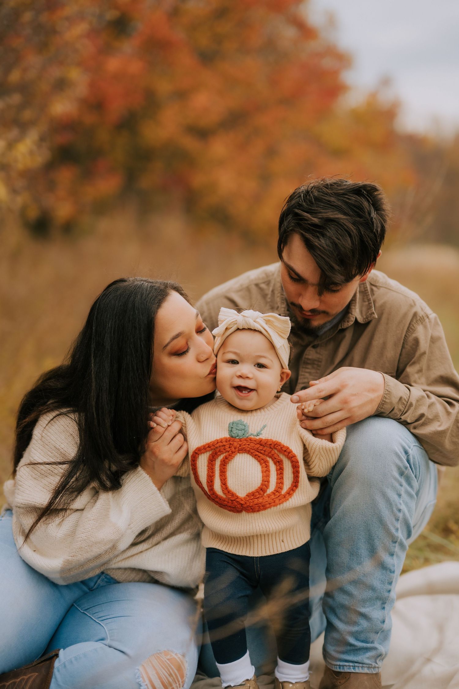 Parents sharing tender moment with baby wearing pumpkin sweater in autumn setting.