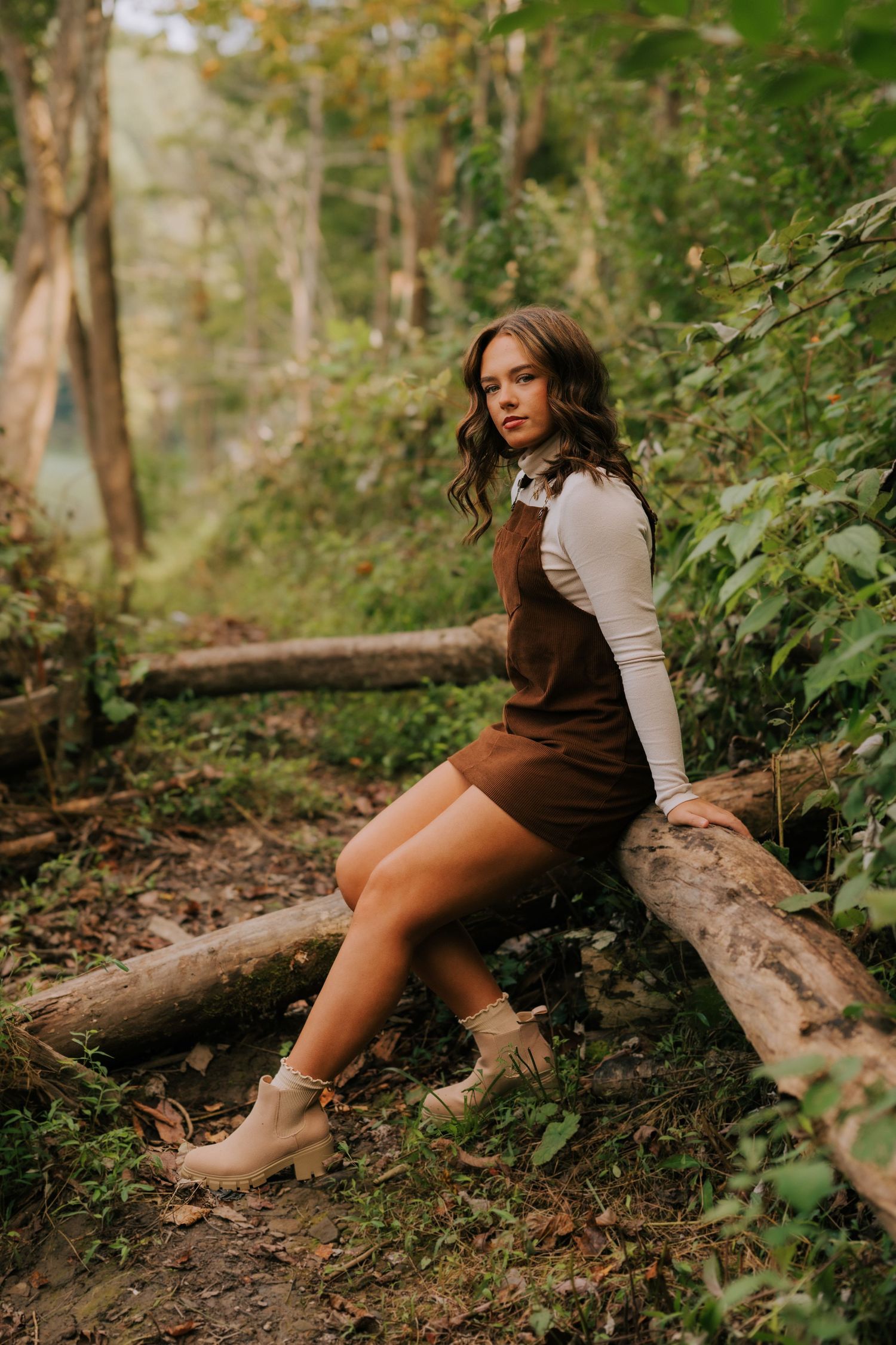 A person in a brown dress and white top poses artistically on fallen logs in a lush forest setting.