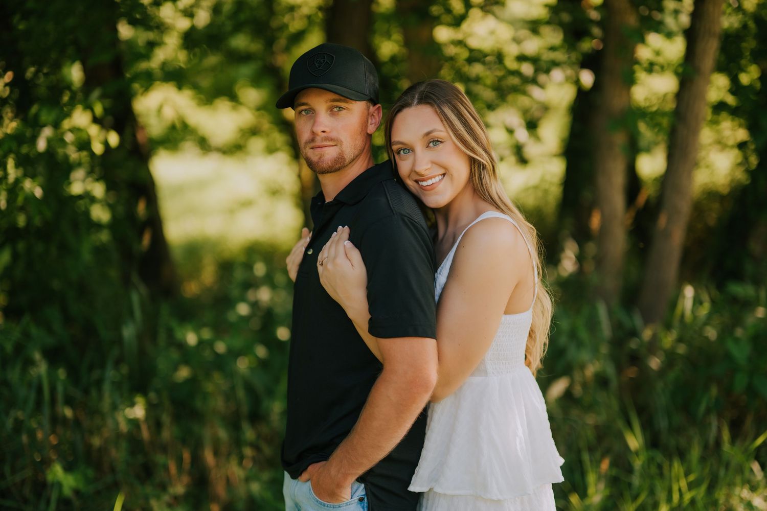 A romantic moment captured between two people in a wooded area with sunlight filtering through trees.