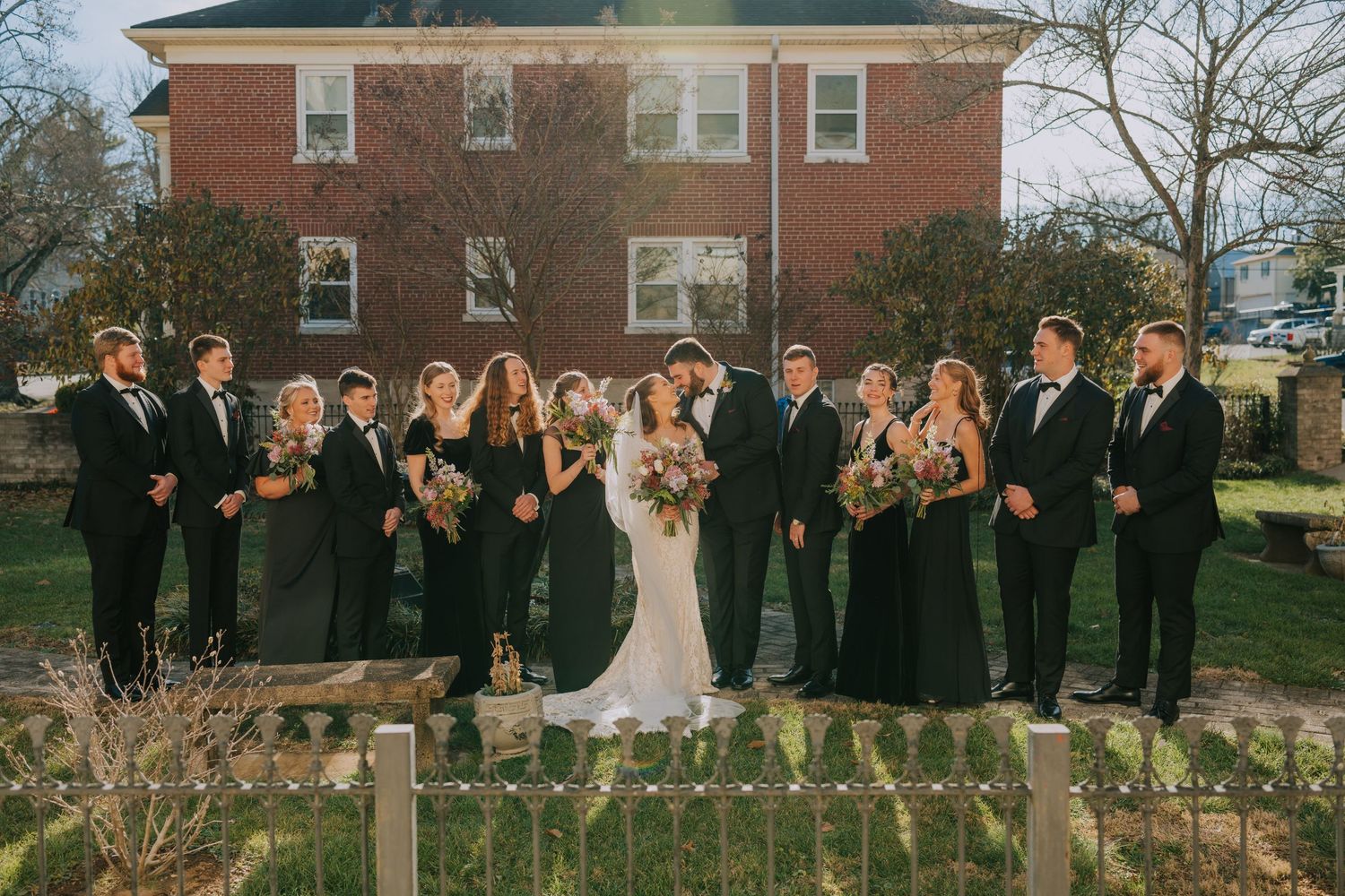 Wedding party wearing black dresses and suits poses together on steps in front of brick building.
