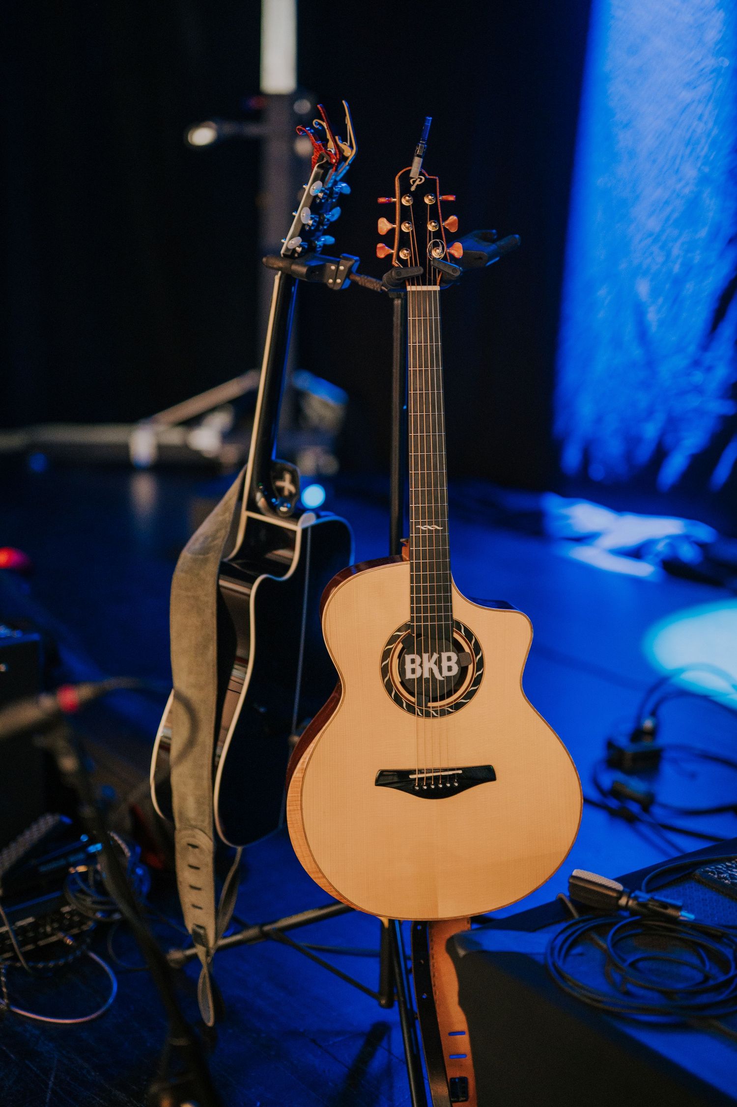 Two acoustic guitars on stage with blue lighting, one light-colored LAG model and one dark, amid performance equipment setup.