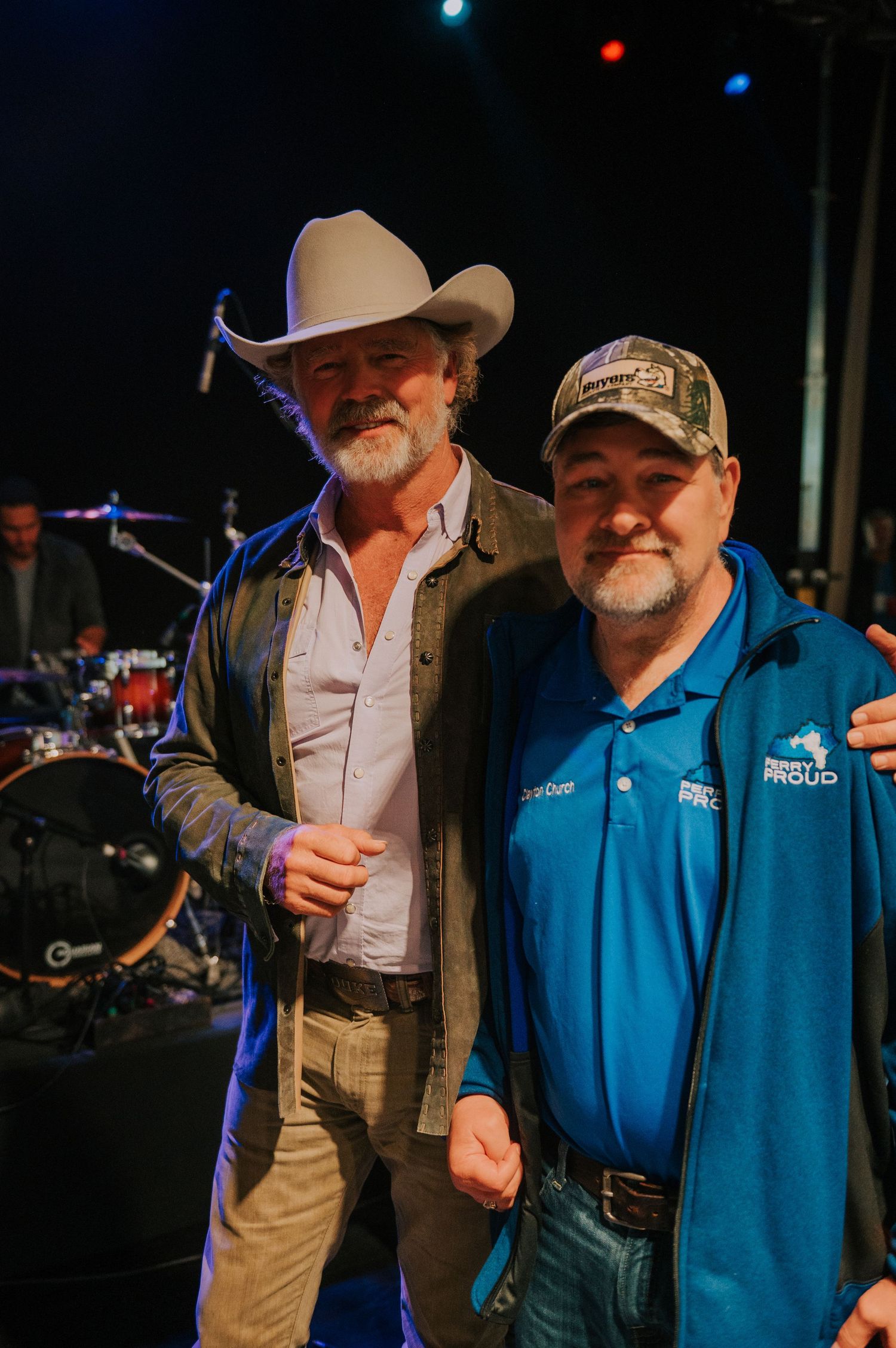 Two men pose together at a concert venue with a drum kit visible in the background.