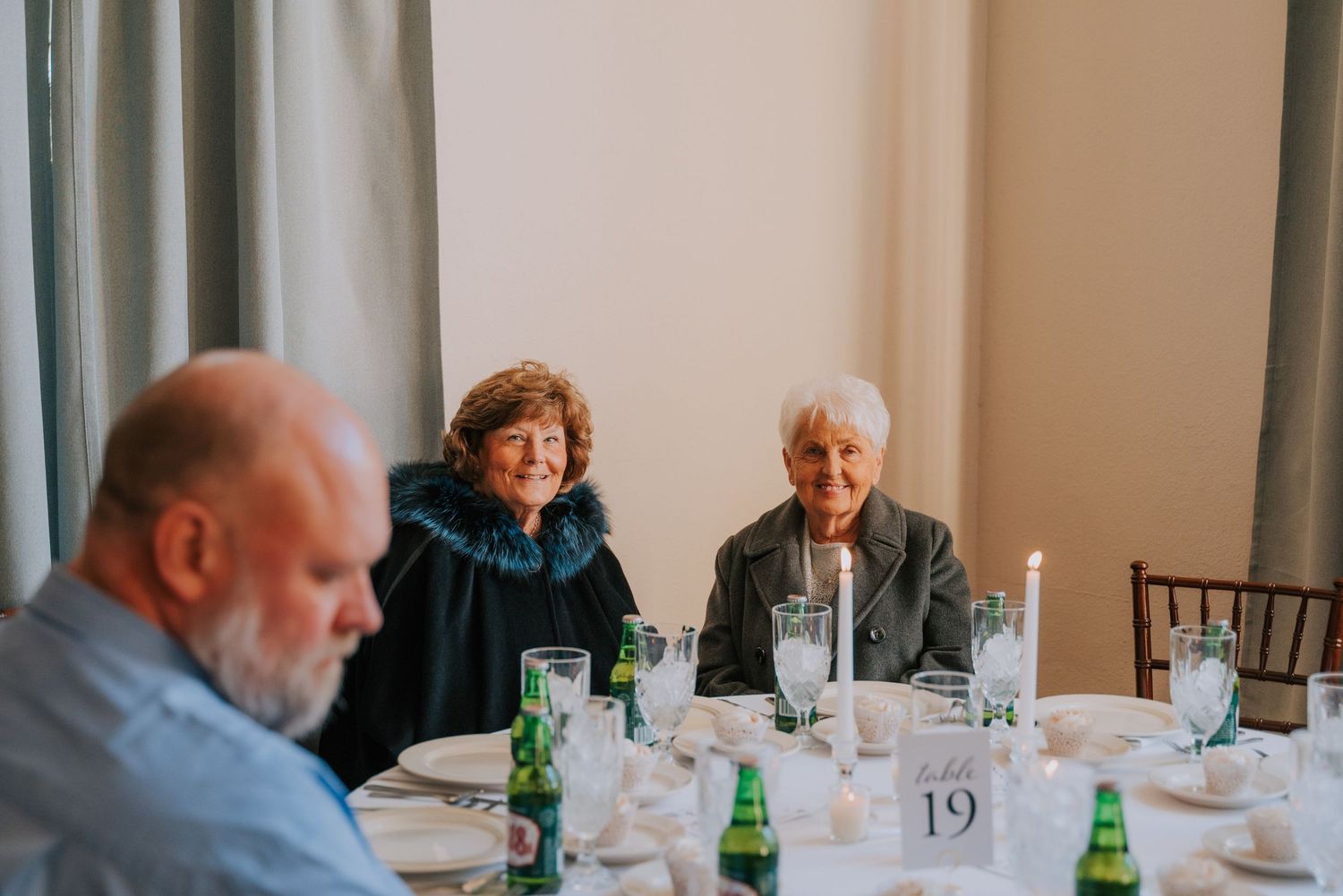 Diners seated at elegant table settings with green glass bottles and numbered centerpieces in a formal event space.