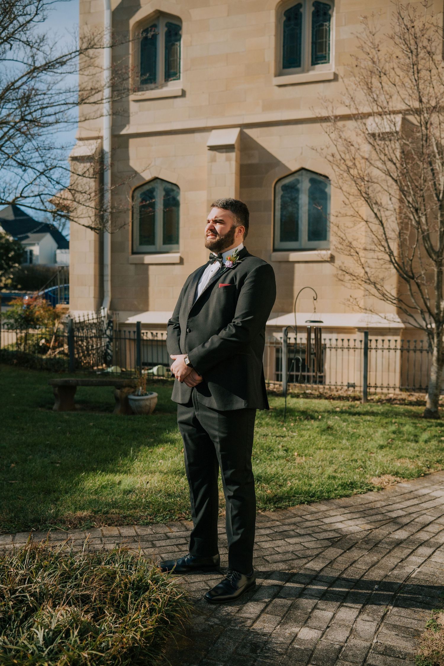 A person stands on a brick pathway outside a beige stone building with arched windows and a green lawn on a sunny day.