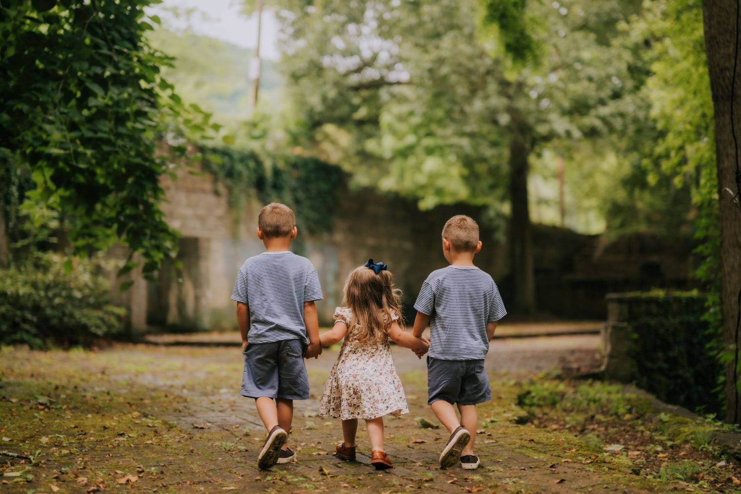 Three young children holding hands walking down a garden path surrounded by lush green trees.