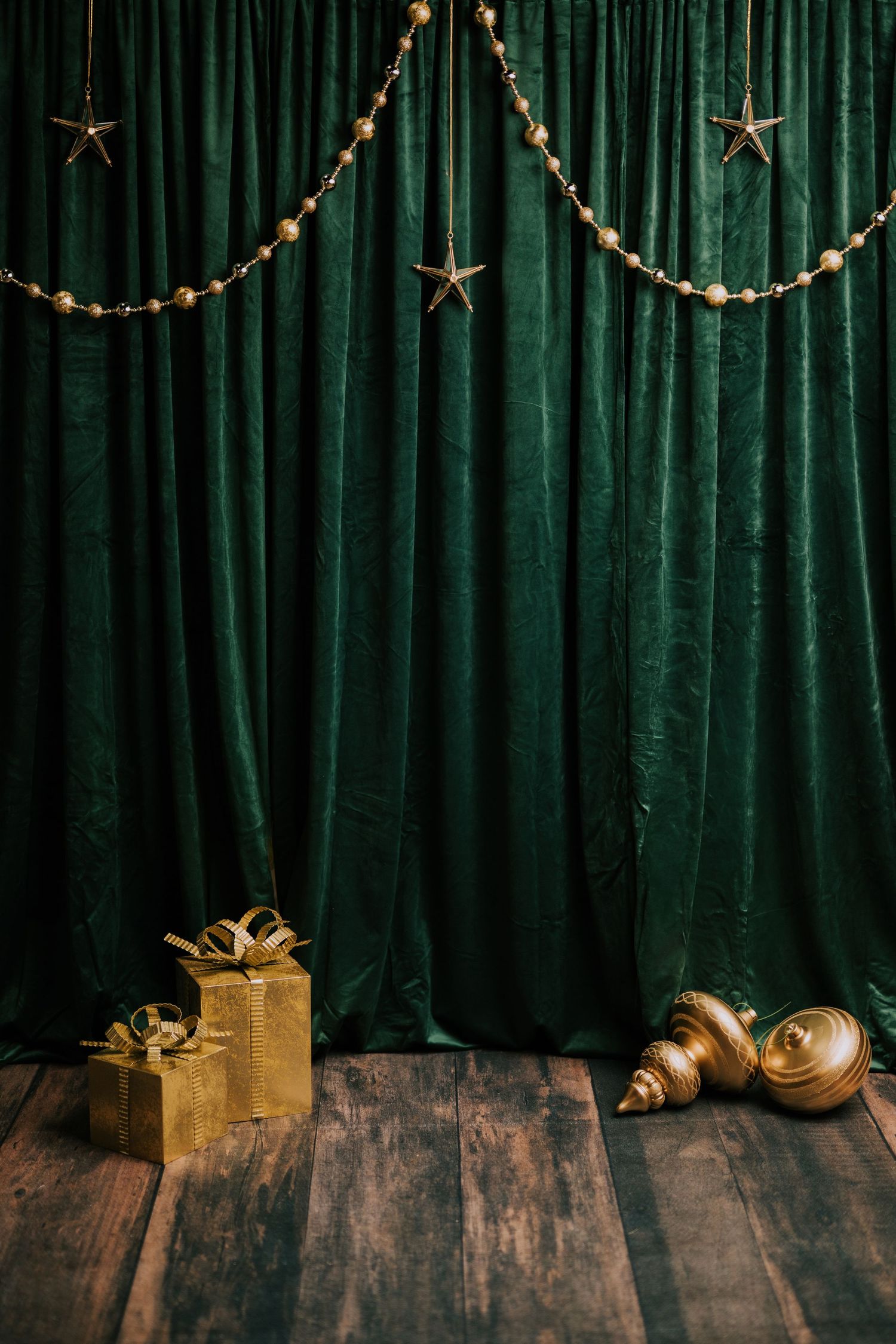 Green velvet curtain backdrop with gold star garland, gift box, and ornaments on rustic wood floor.