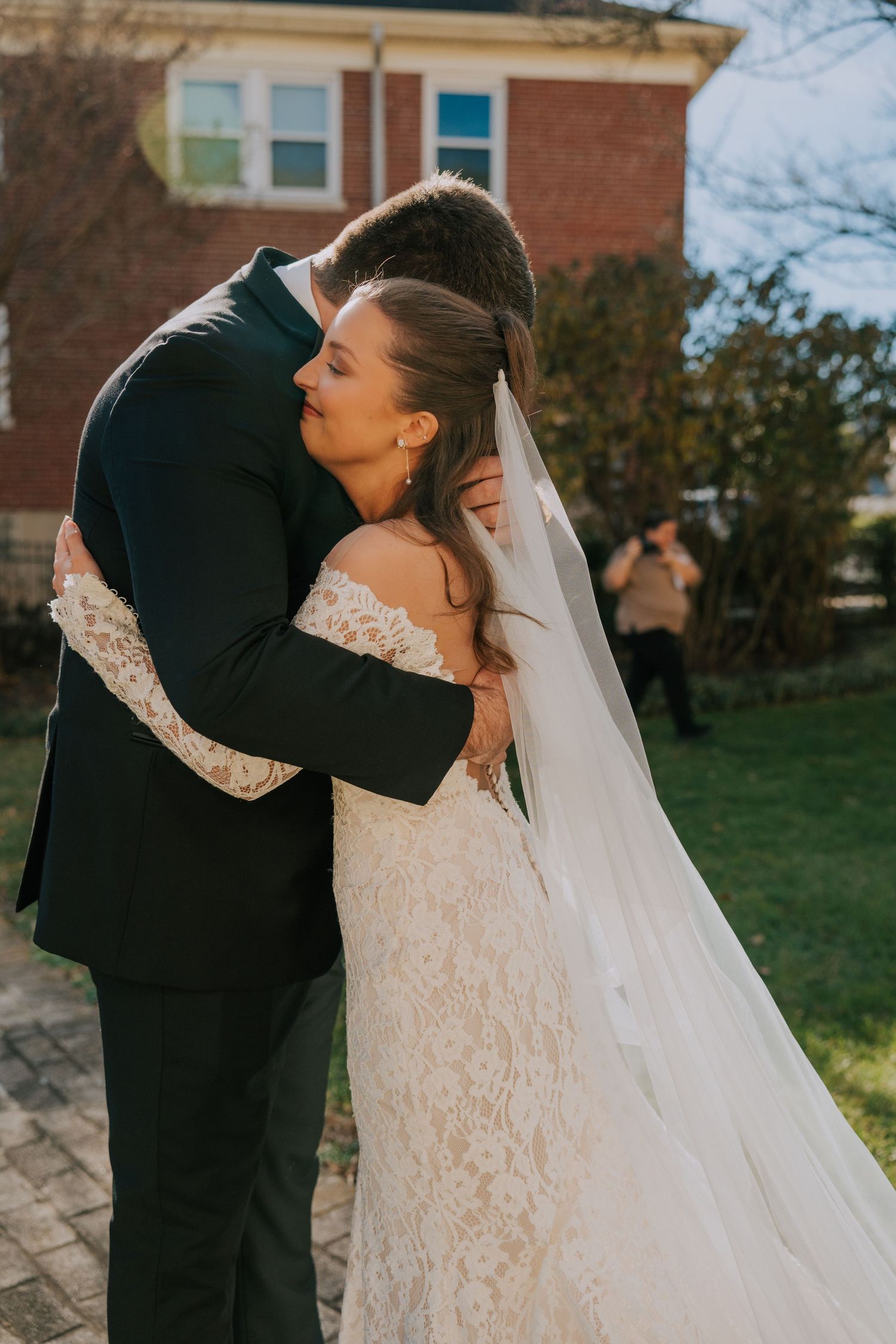 An intimate wedding moment captured outdoors with a couple embracing in front of a brick building on a sunny day.