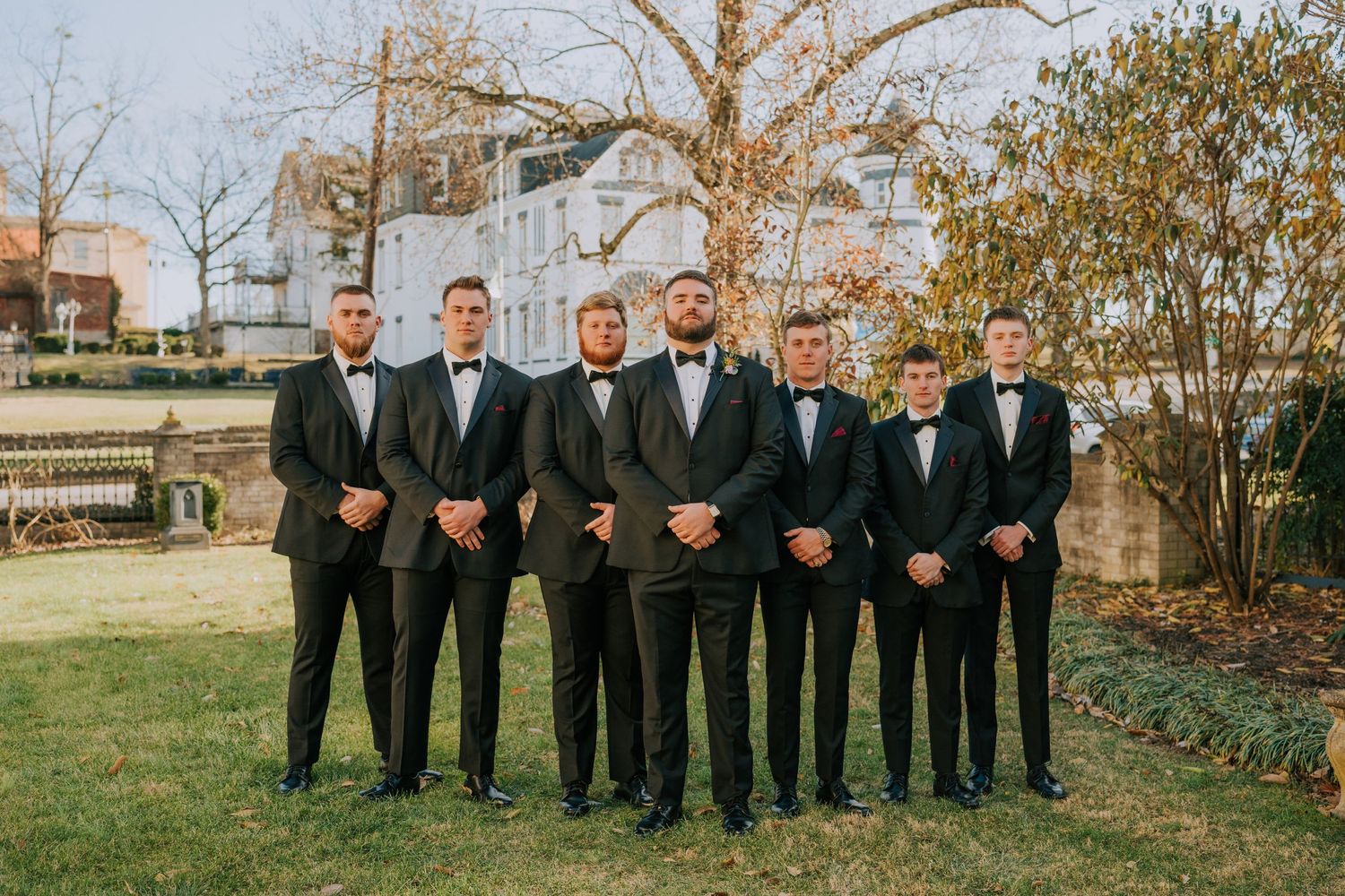 A group in black tuxedos and bowties stand together on a lawn for a formal wedding party photo.