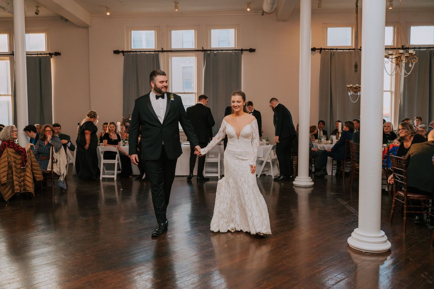 A couple walks across an elegant reception venue with white columns and a polished dark floor while guests look on.