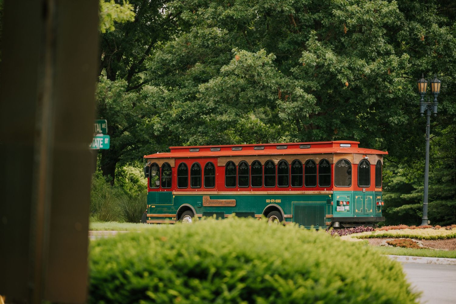 A vintage red trolley bus with arched windows travels through a lush green park setting.