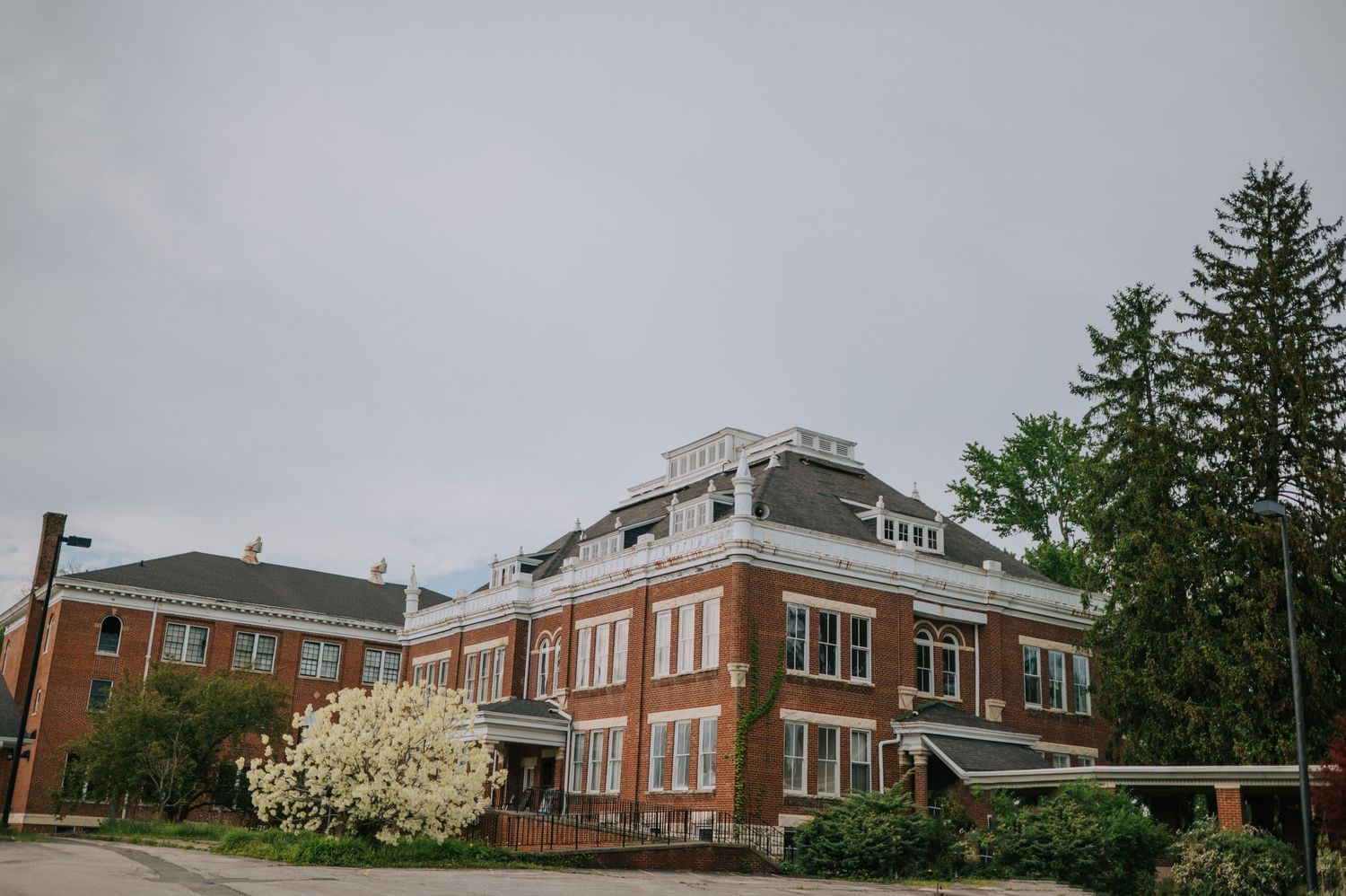 A historic brick building with white trim and flowering trees in bloom against an overcast sky.