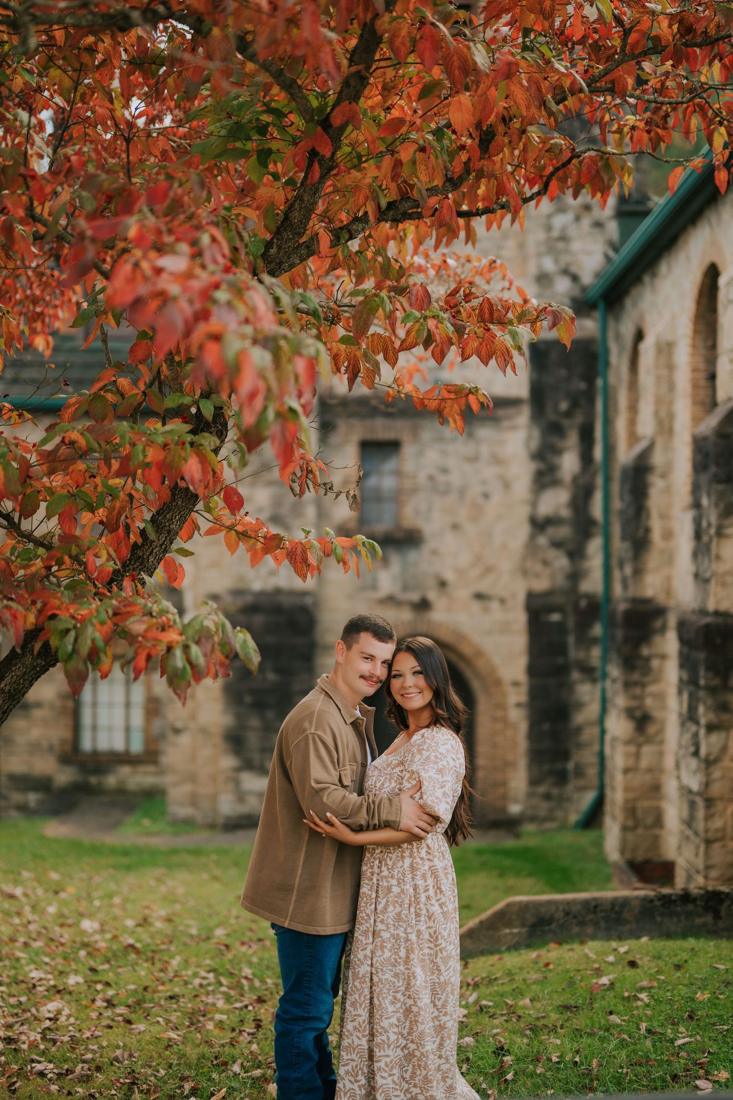 A couple shares an intimate moment beneath vibrant autumn foliage near a stone building.