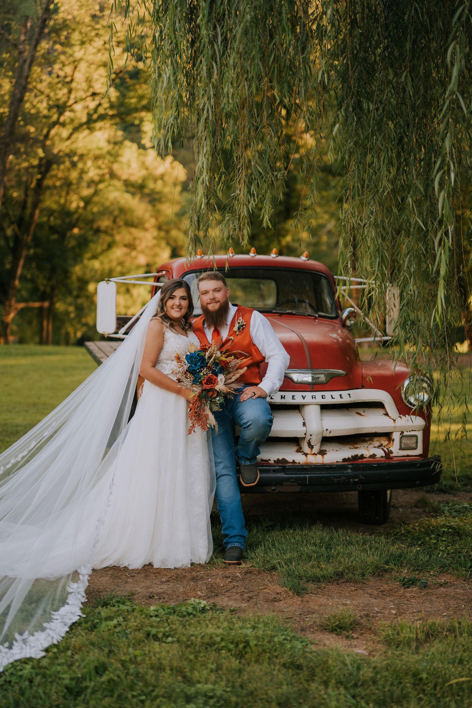 A newlywed couple poses in front of a vintage red Chevrolet truck under willow trees.