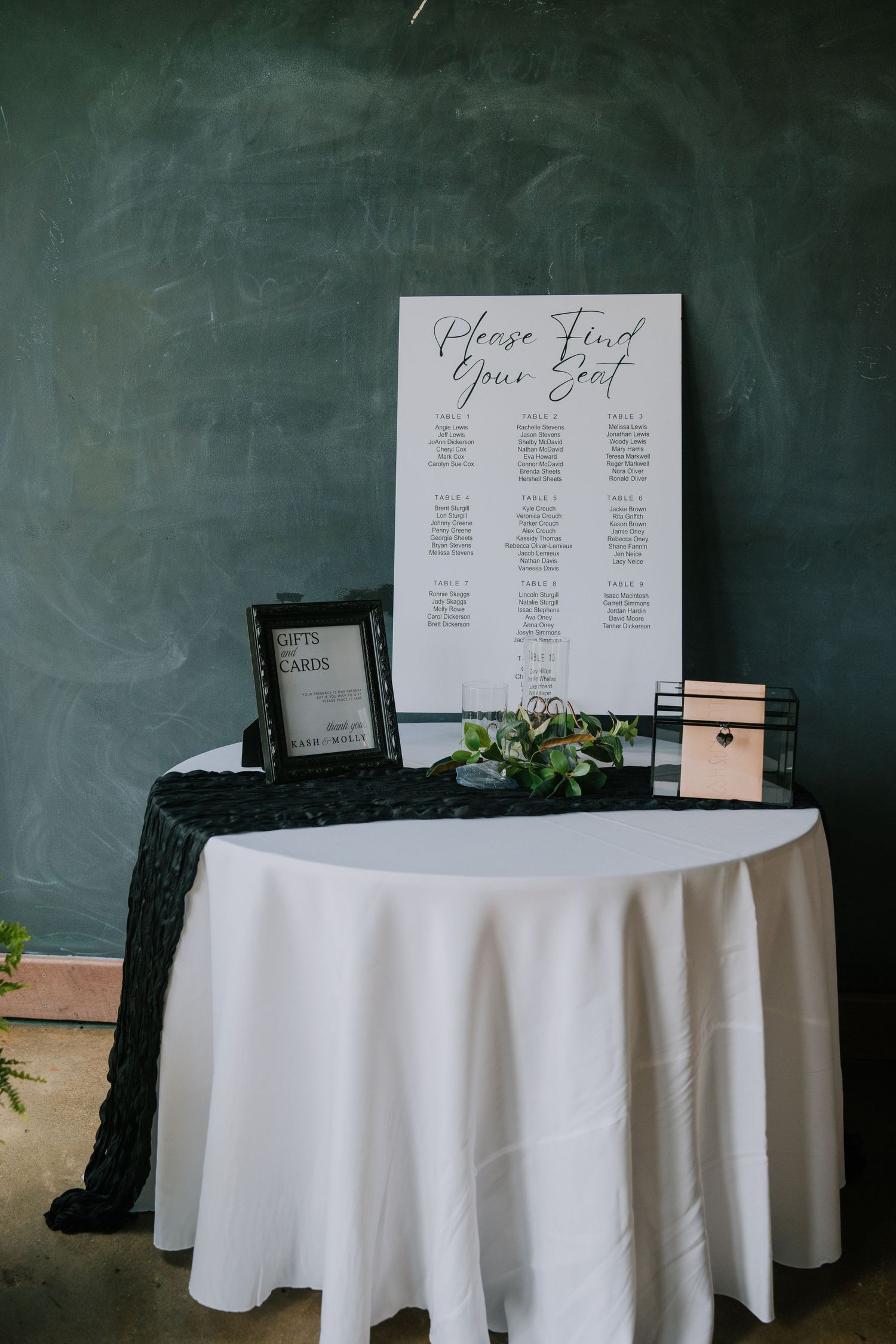 Wedding reception seating chart display with white tablecloths and chalkboard backdrop.