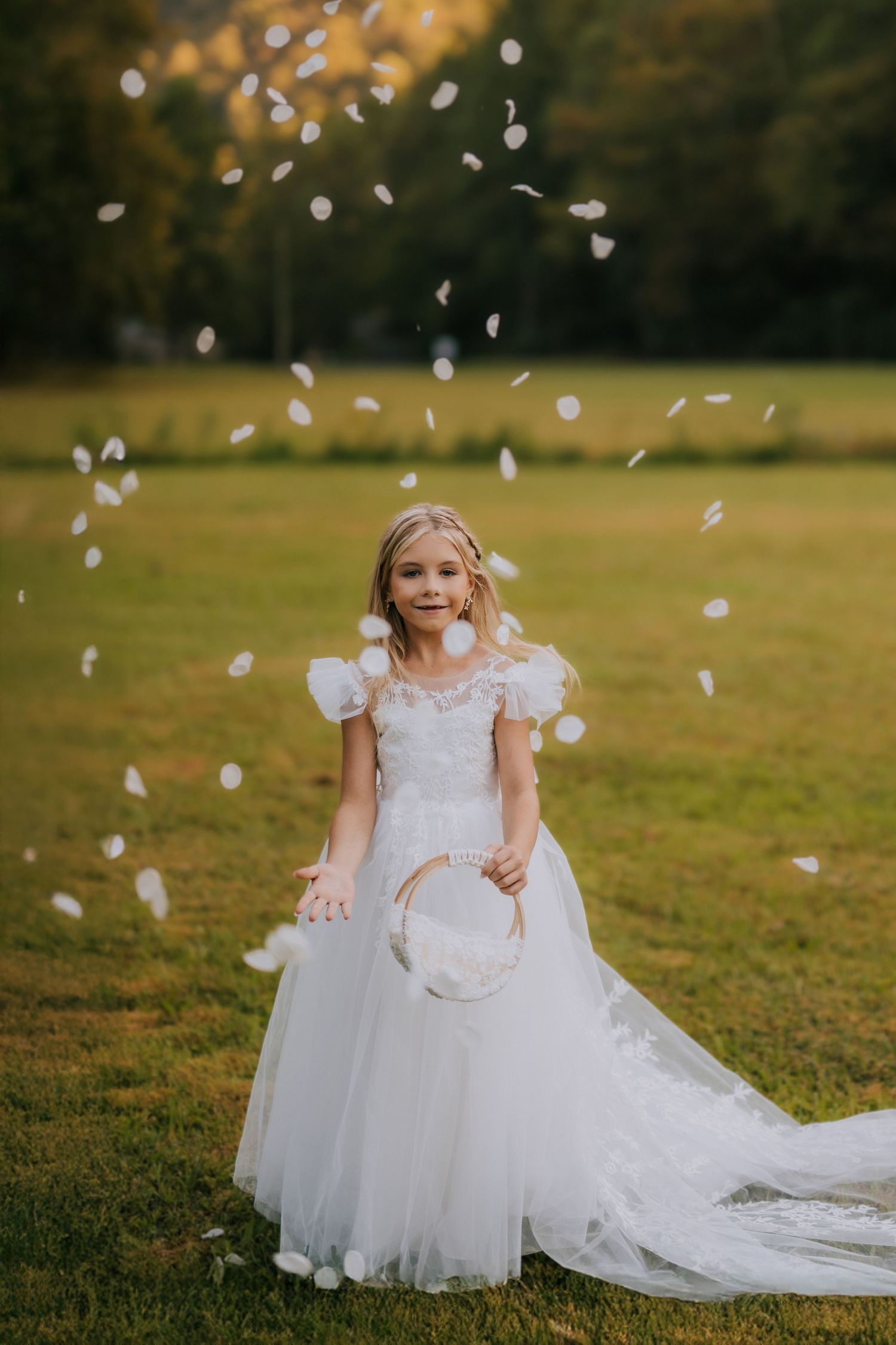 Young flower girl in white dress playing with confetti in a sunlit meadow.