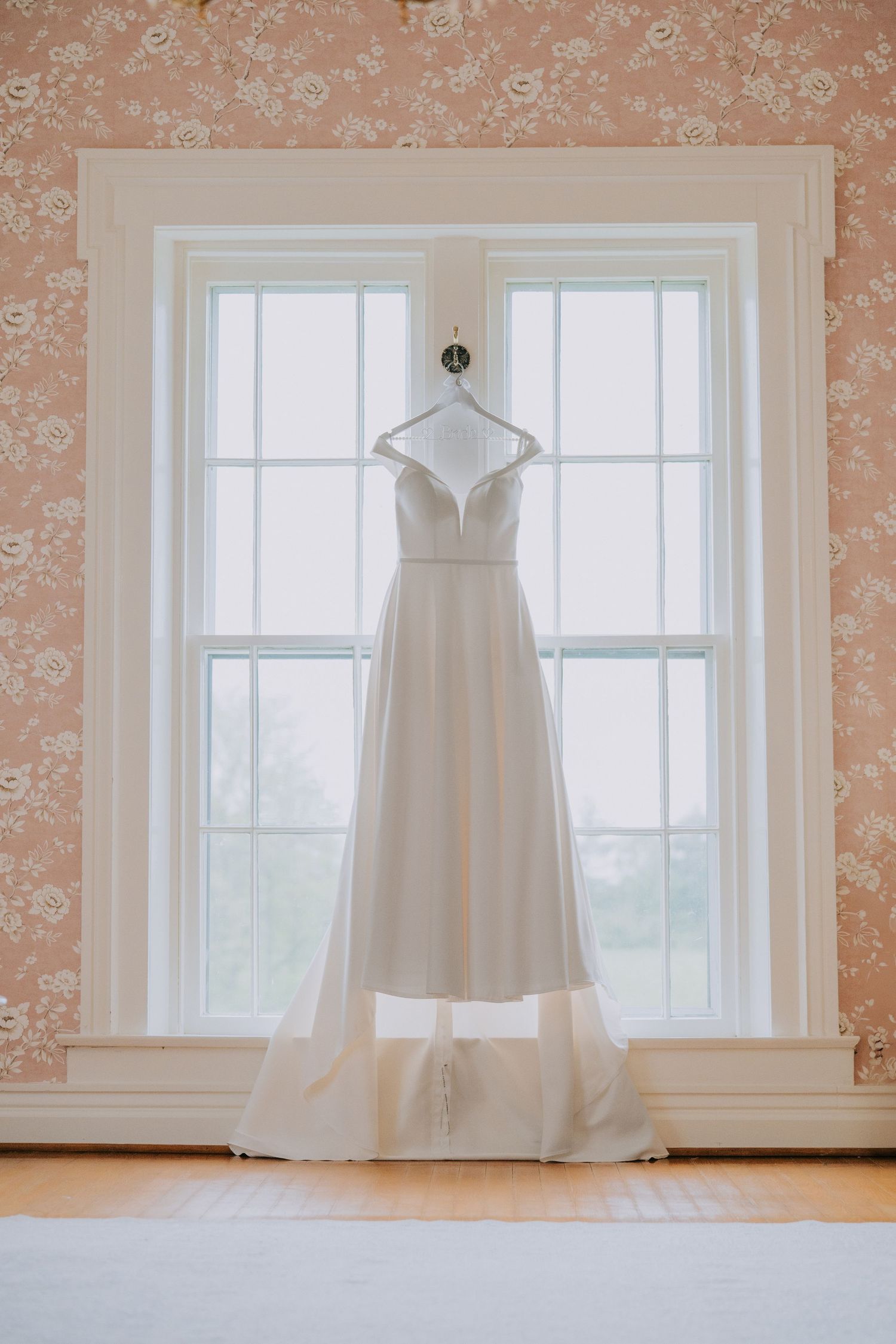 A classic white wedding dress hangs elegantly against pink floral wallpaper near bright windows.