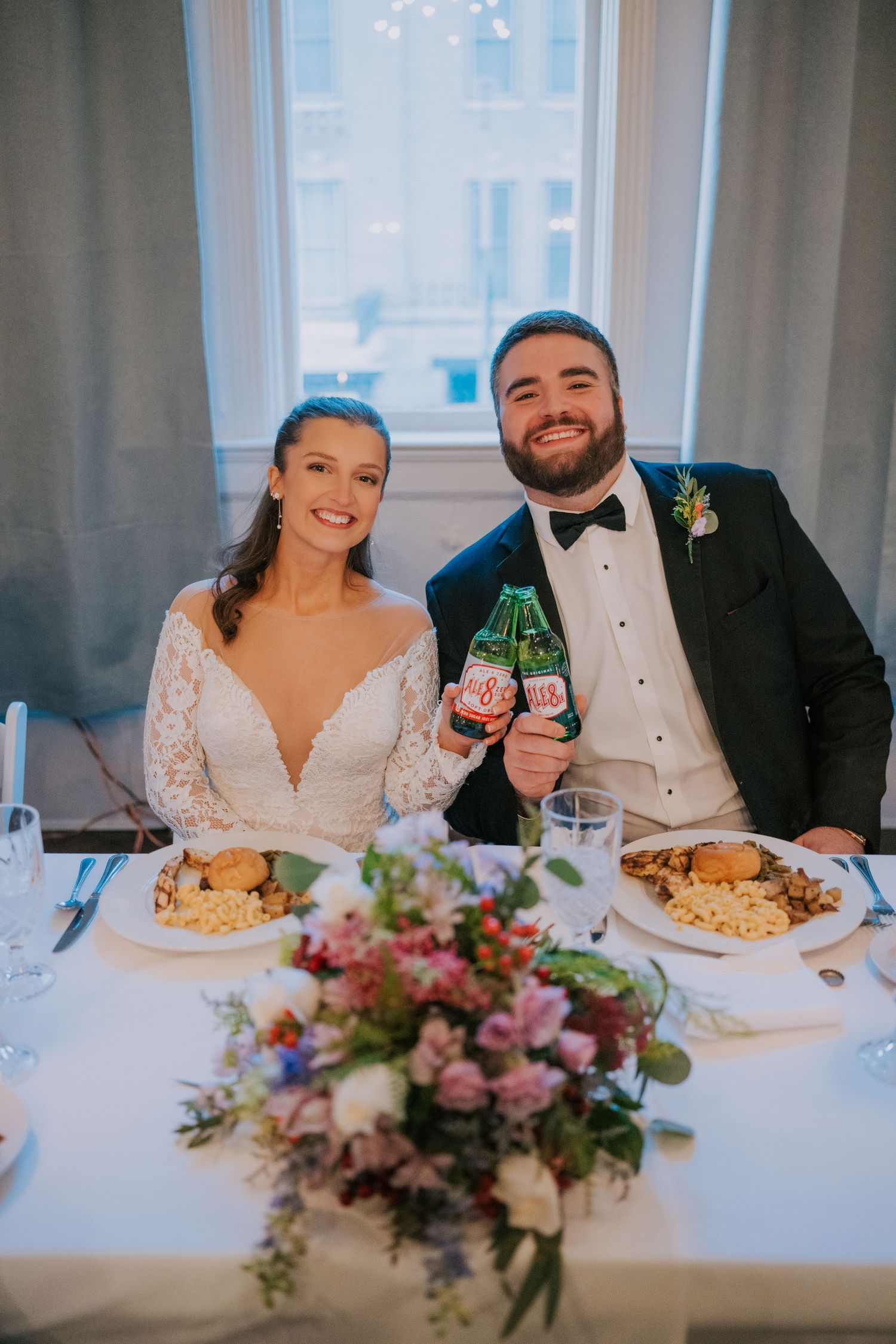 Newlyweds toast with drinks at reception dinner table decorated with pink floral centerpiece.
