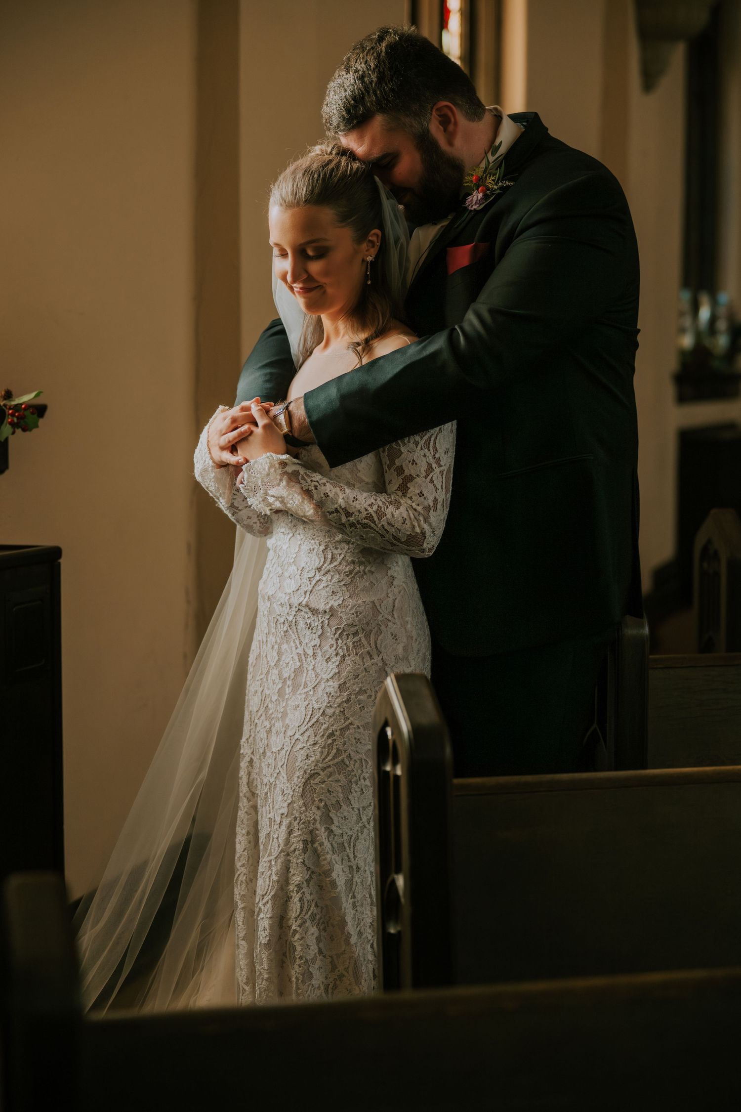 A romantic wedding moment captured in soft lighting as a couple embraces in front of a church window.