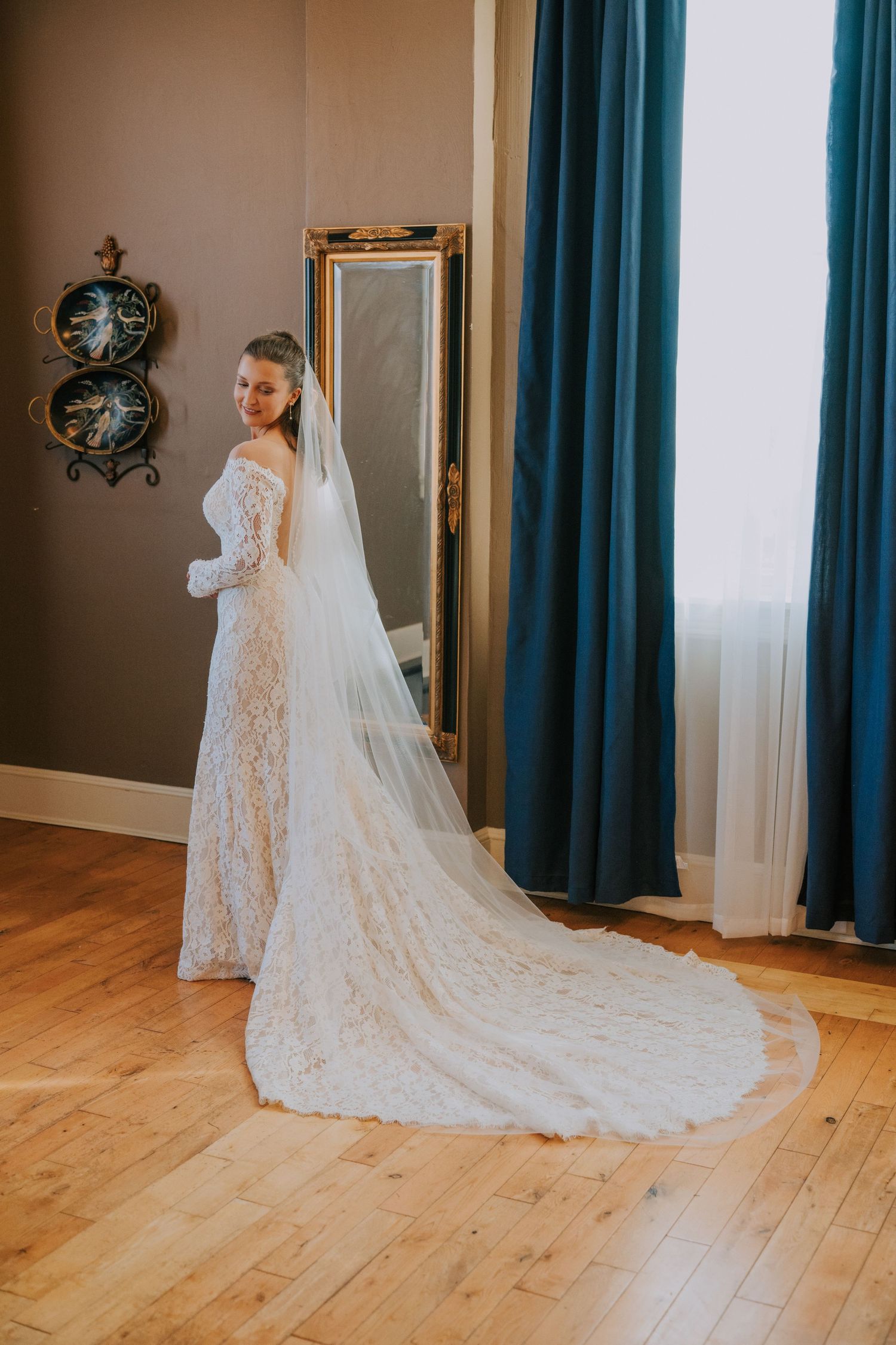 A bride in a long lace wedding gown with cathedral veil poses in a room with teal curtains and hardwood floors.
