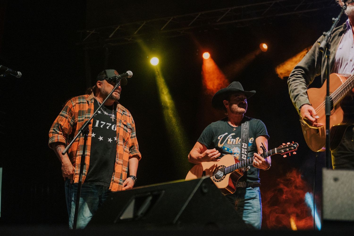Country music performers on stage with acoustic guitars under colorful stage lighting at night during a live concert.