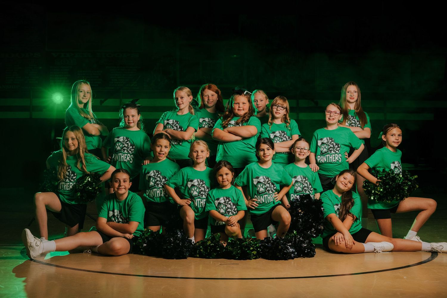 Group photo of cheerleaders in green uniforms posing together on a dark stage with dramatic lighting.