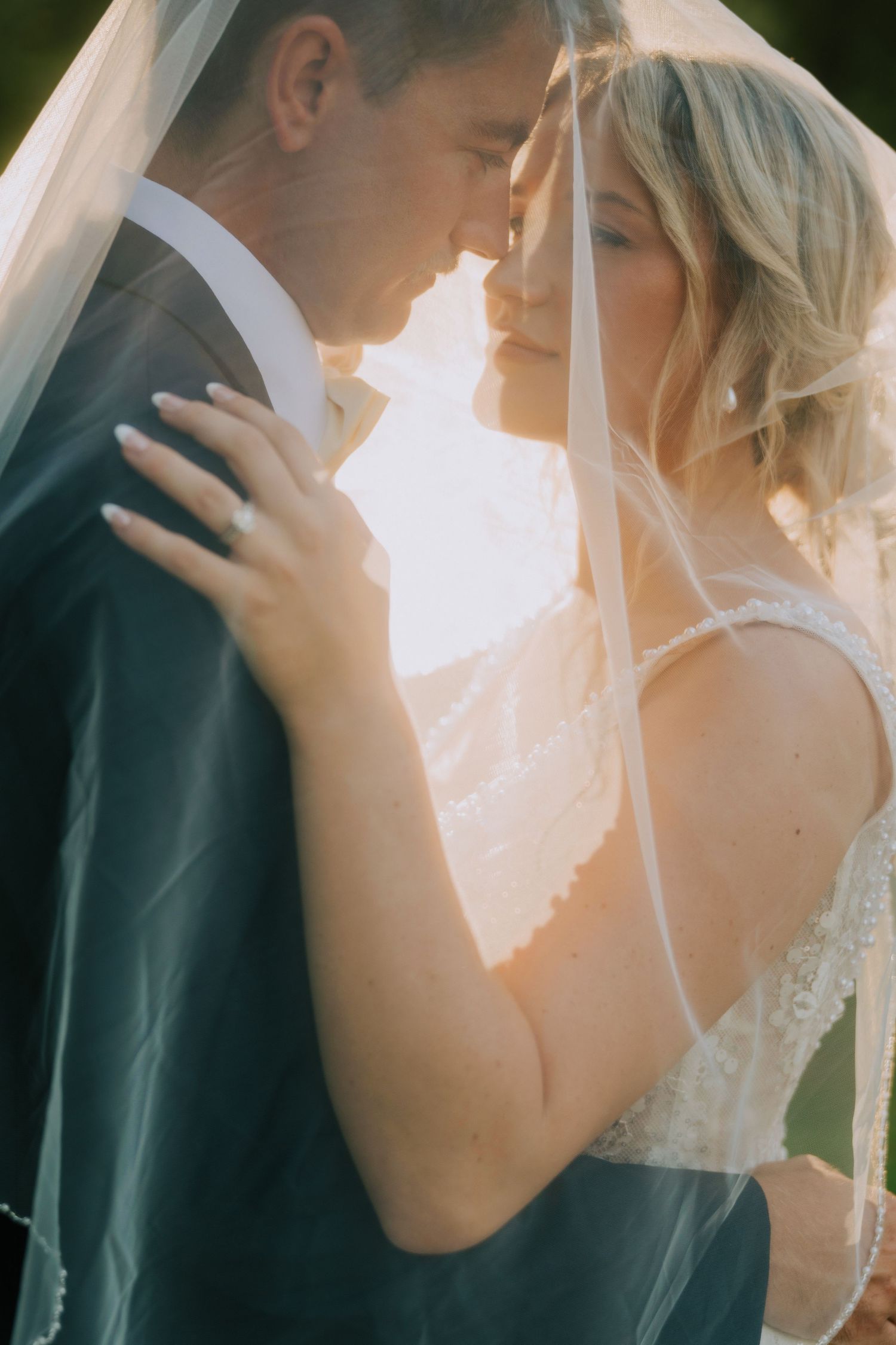 Bride and groom share an intimate moment under a flowing veil, bathed in warm golden sunset light.