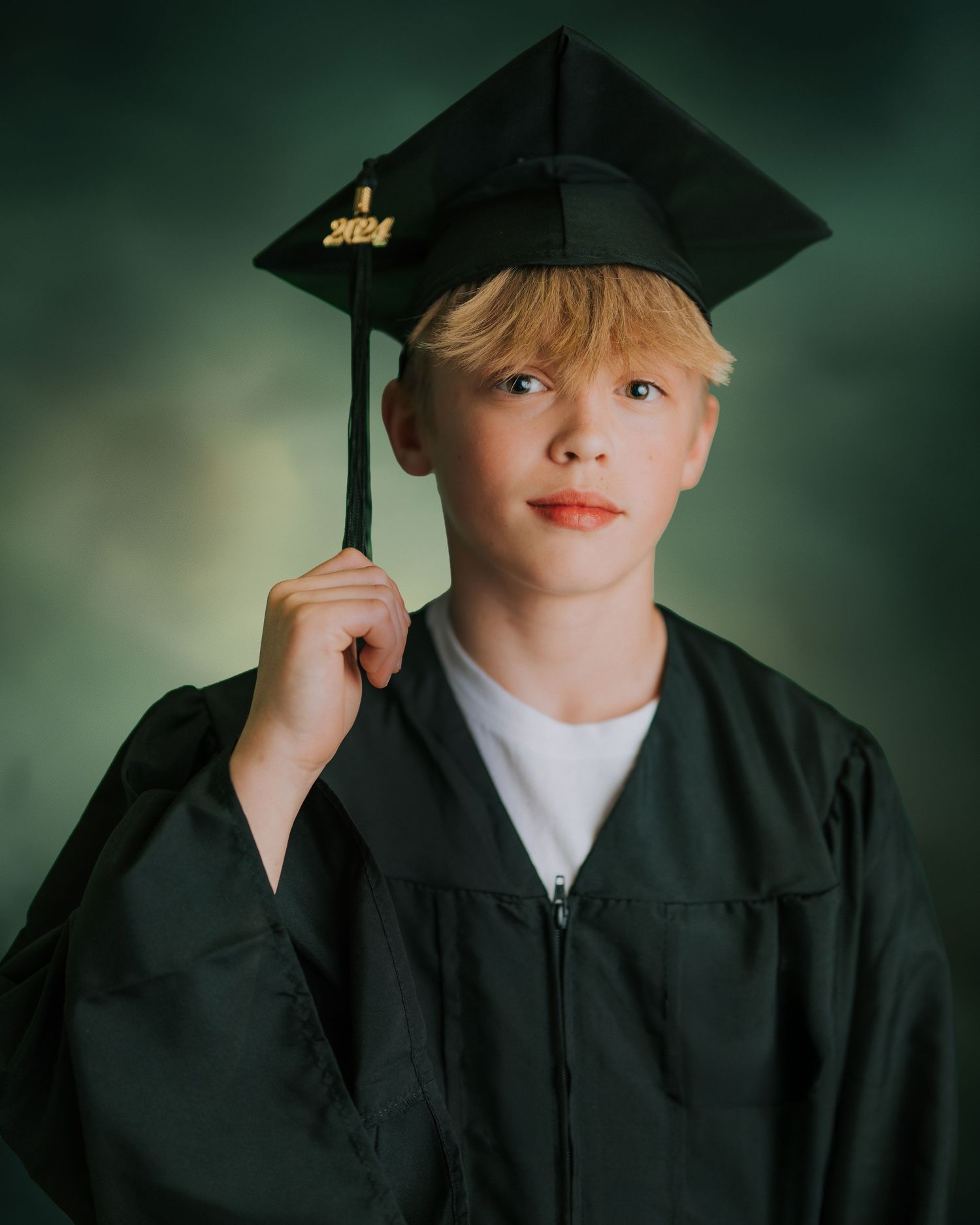 Young boy in dark green graduation cap and gown holding tassel, class of 2024 graduate portrait photo.