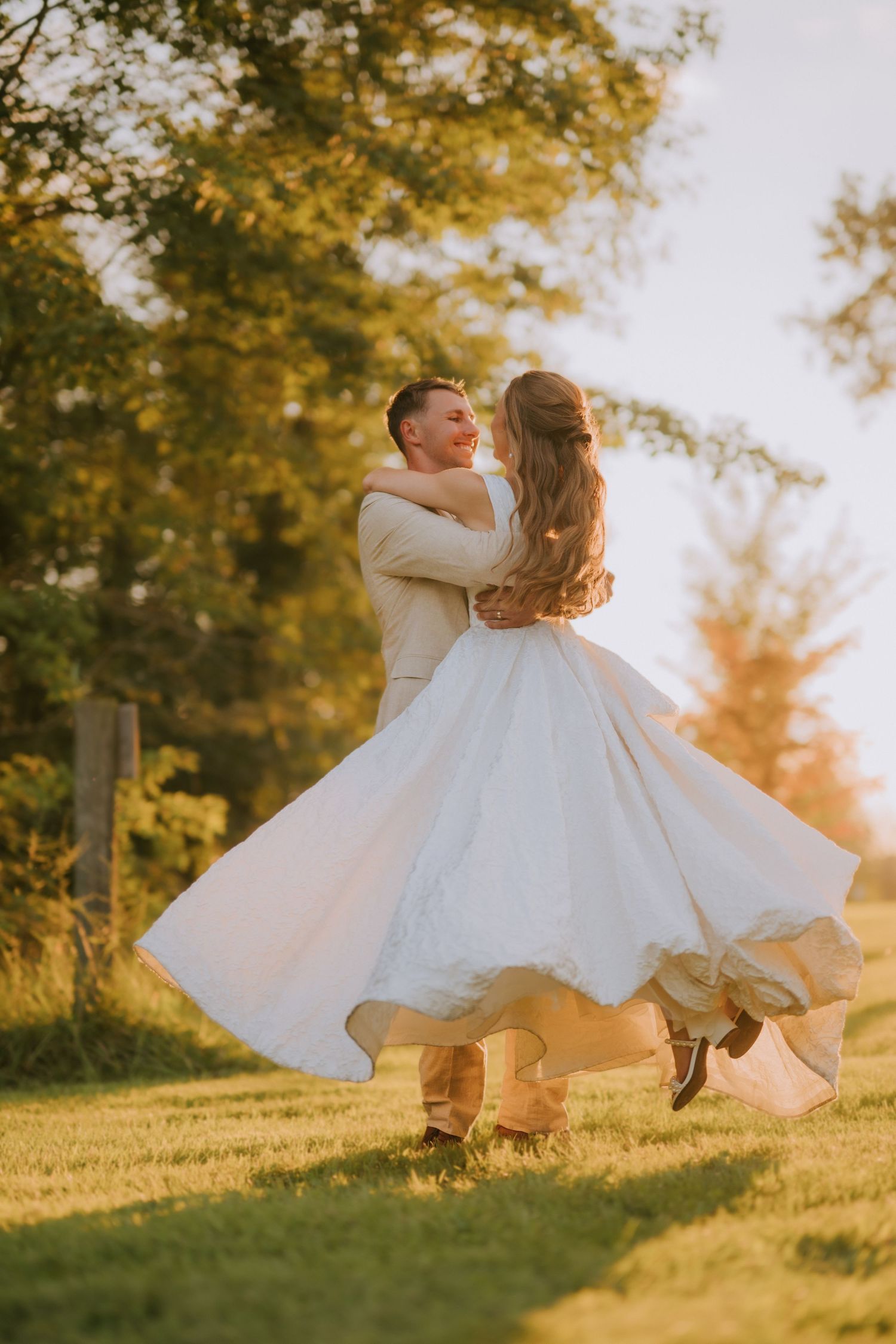 Groom lifting bride in white and gold dress during golden hour outdoor wedding photo on green lawn.
