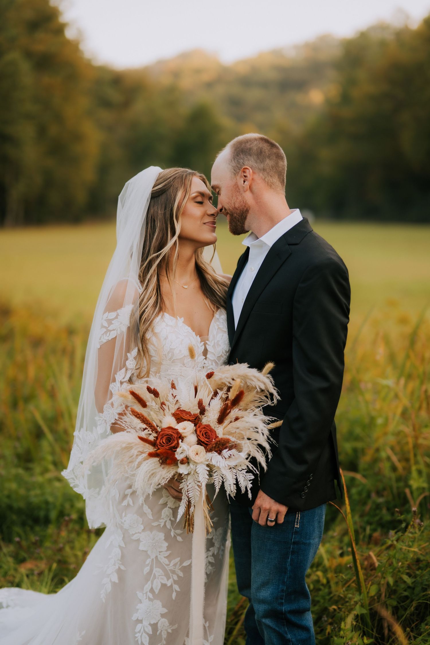 A romantic wedding couple stands in a golden field at sunset holding a dried floral bouquet.