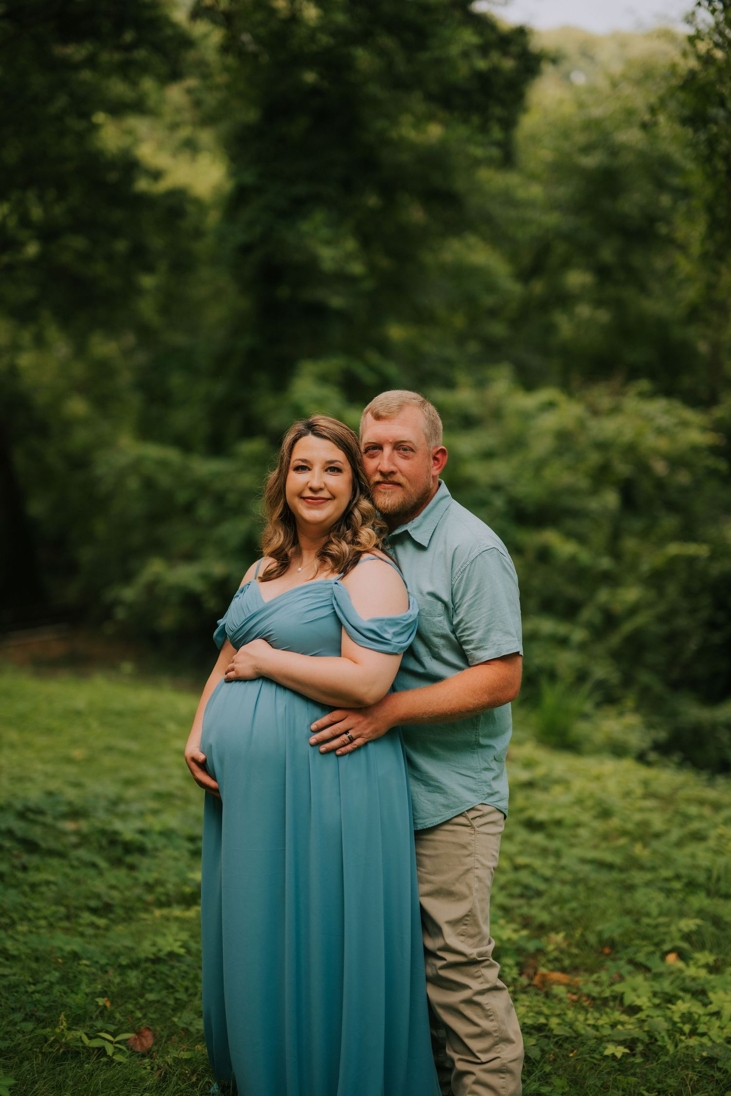 Romantic maternity photos in a green garden setting with couple in casual attire.