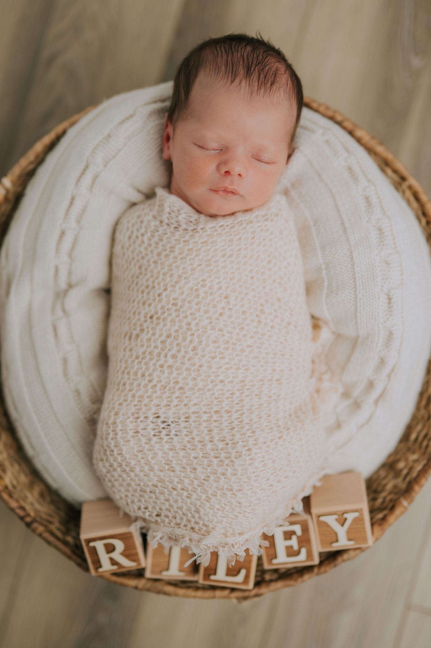 A newborn wrapped in white knit blanket sleeping in basket with wooden letter blocks.