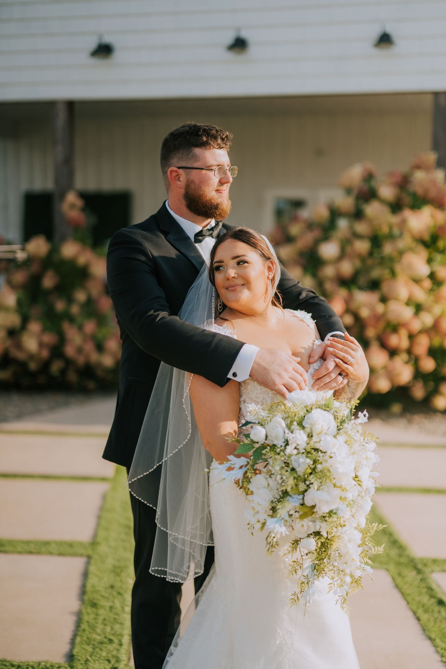A newlywed couple embraces outdoors while holding a cascading white floral bouquet.