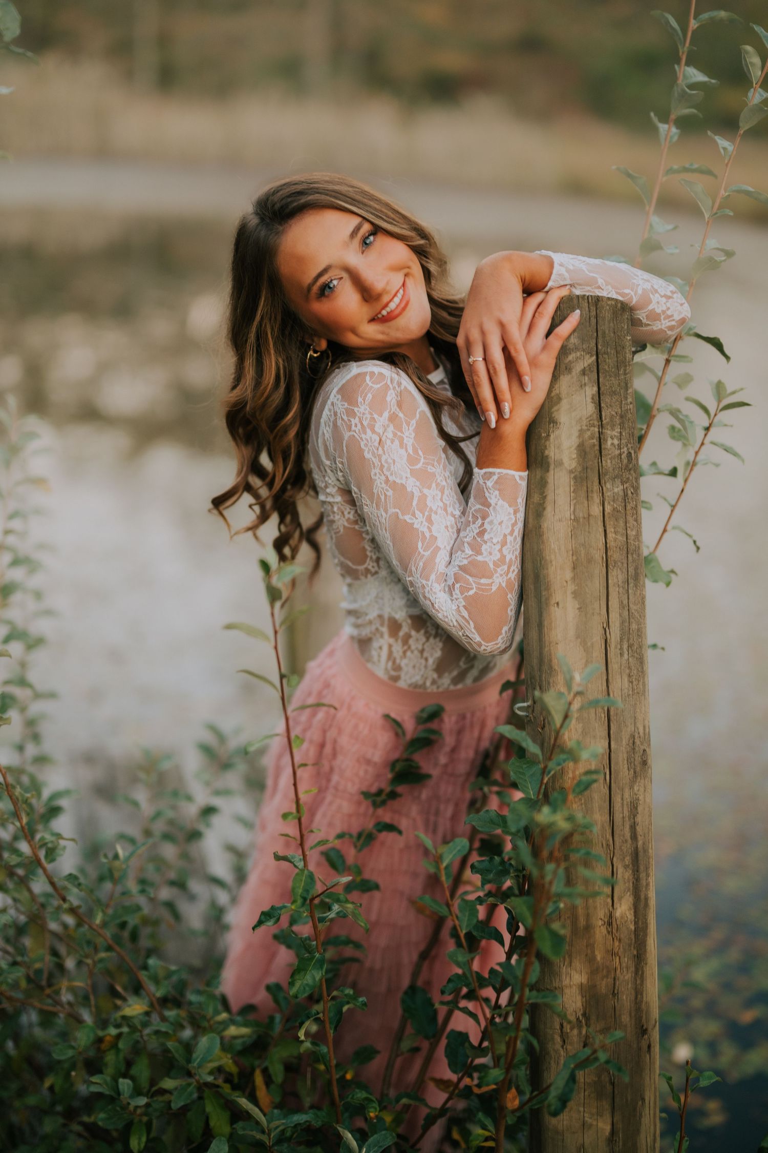 Romantic portrait series by water featuring lace dress and wooden fence post.