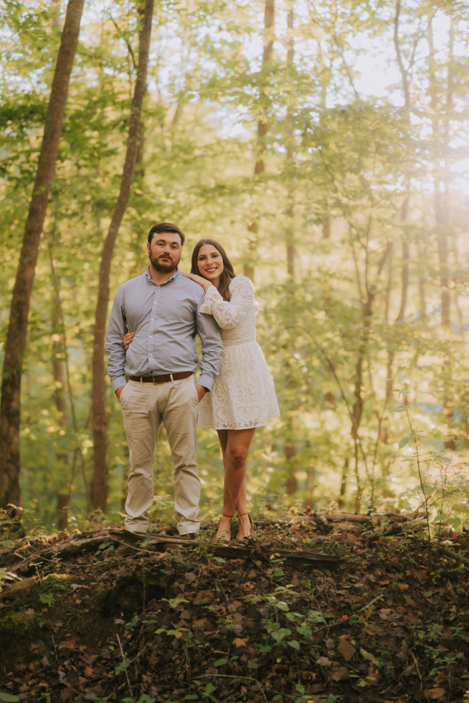 Couple poses together on a wooded hillside with sunlit trees in the background.