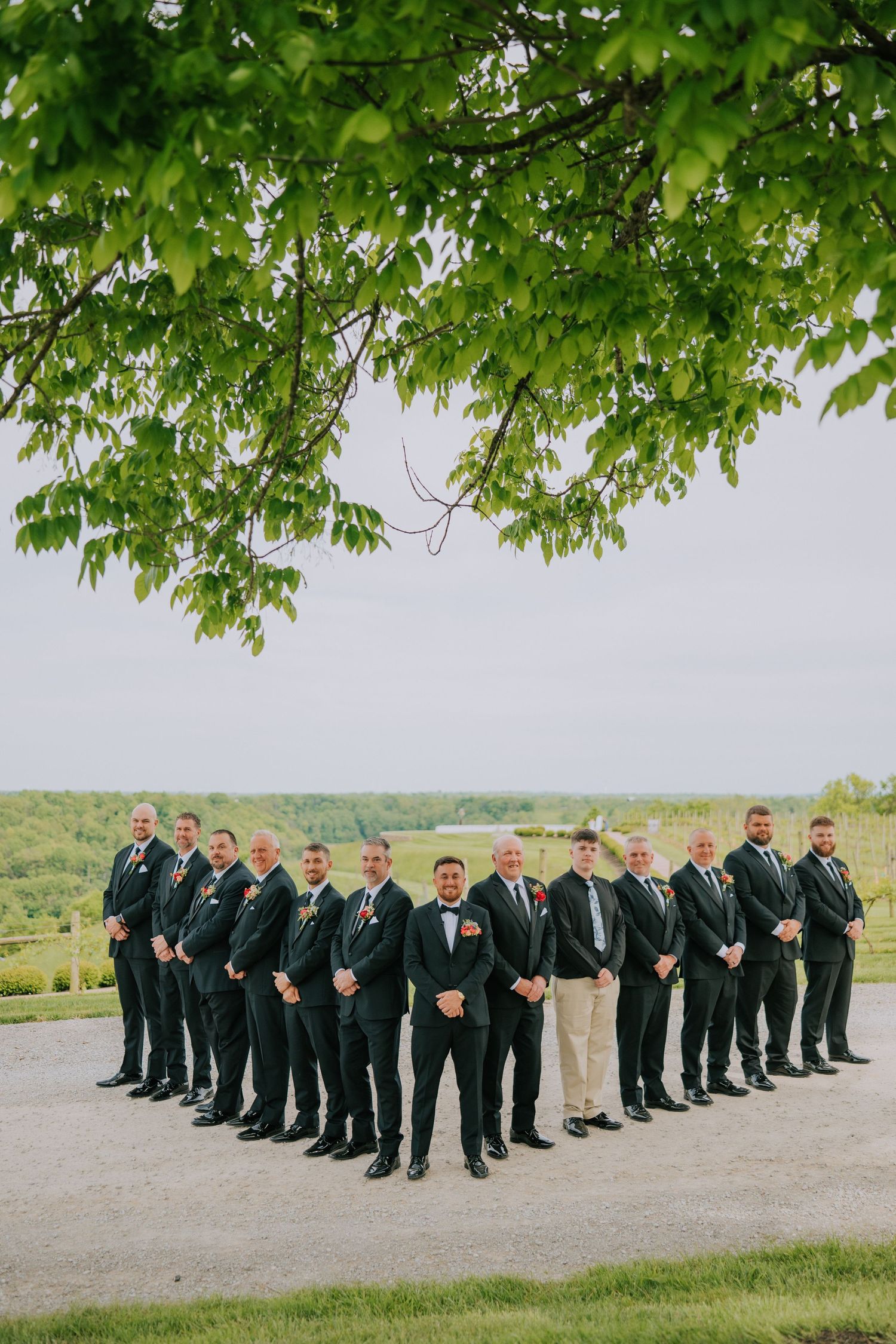 Large groomsmen party lined up outdoors under green tree canopy wearing dark suits.