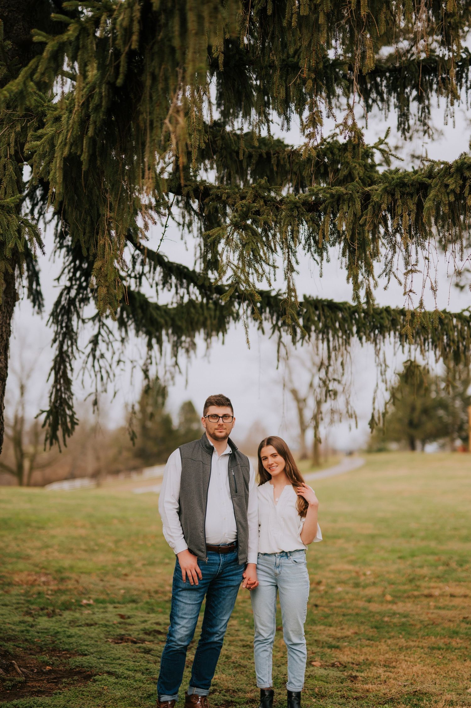 A couple poses together under a large evergreen tree on a grassy hill during an outdoor photoshoot.