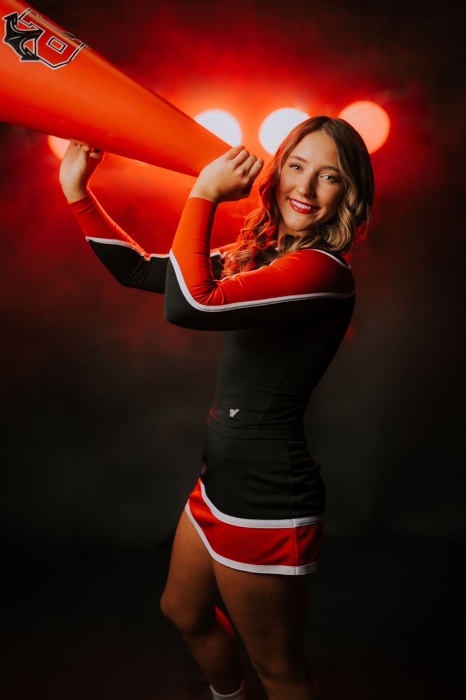 Cheerleader in black and red uniform poses with orange megaphone against dark background with red lighting effects.