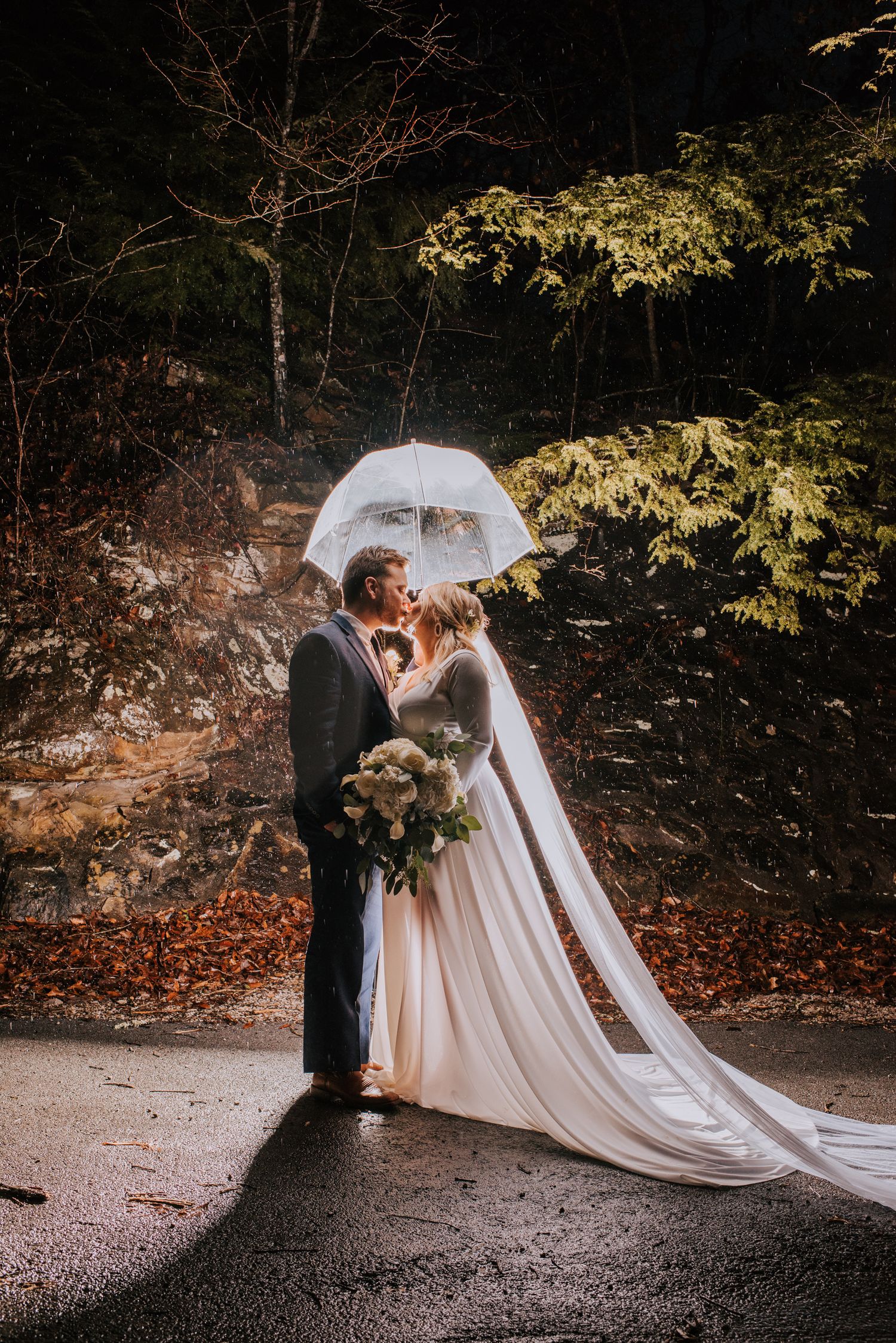 A romantic wedding couple poses under a white umbrella in a wooded forest setting with autumn leaves.
