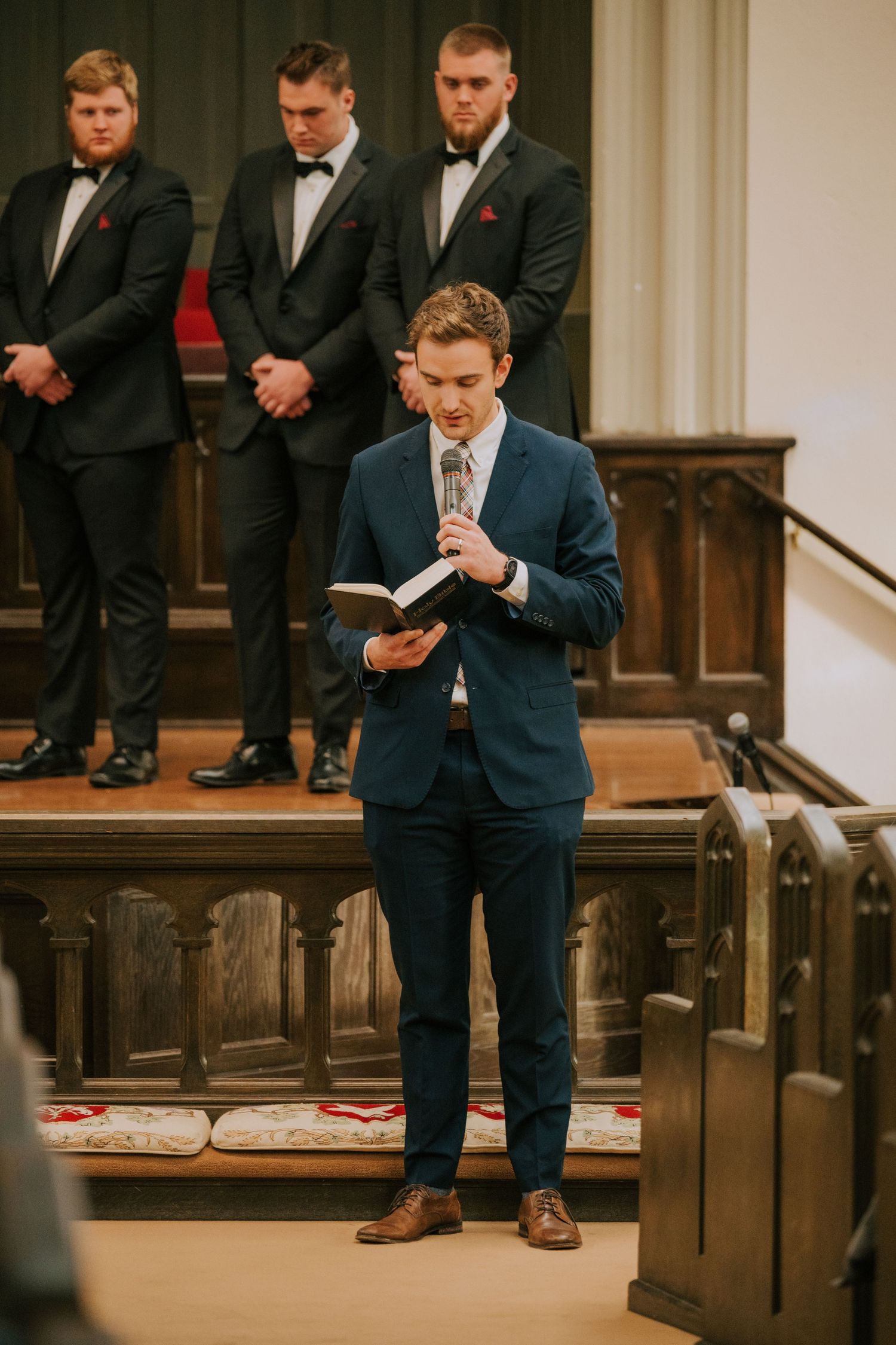 A speaker in a navy suit stands at a church podium while groomsmen in black tuxedos look on from behind.