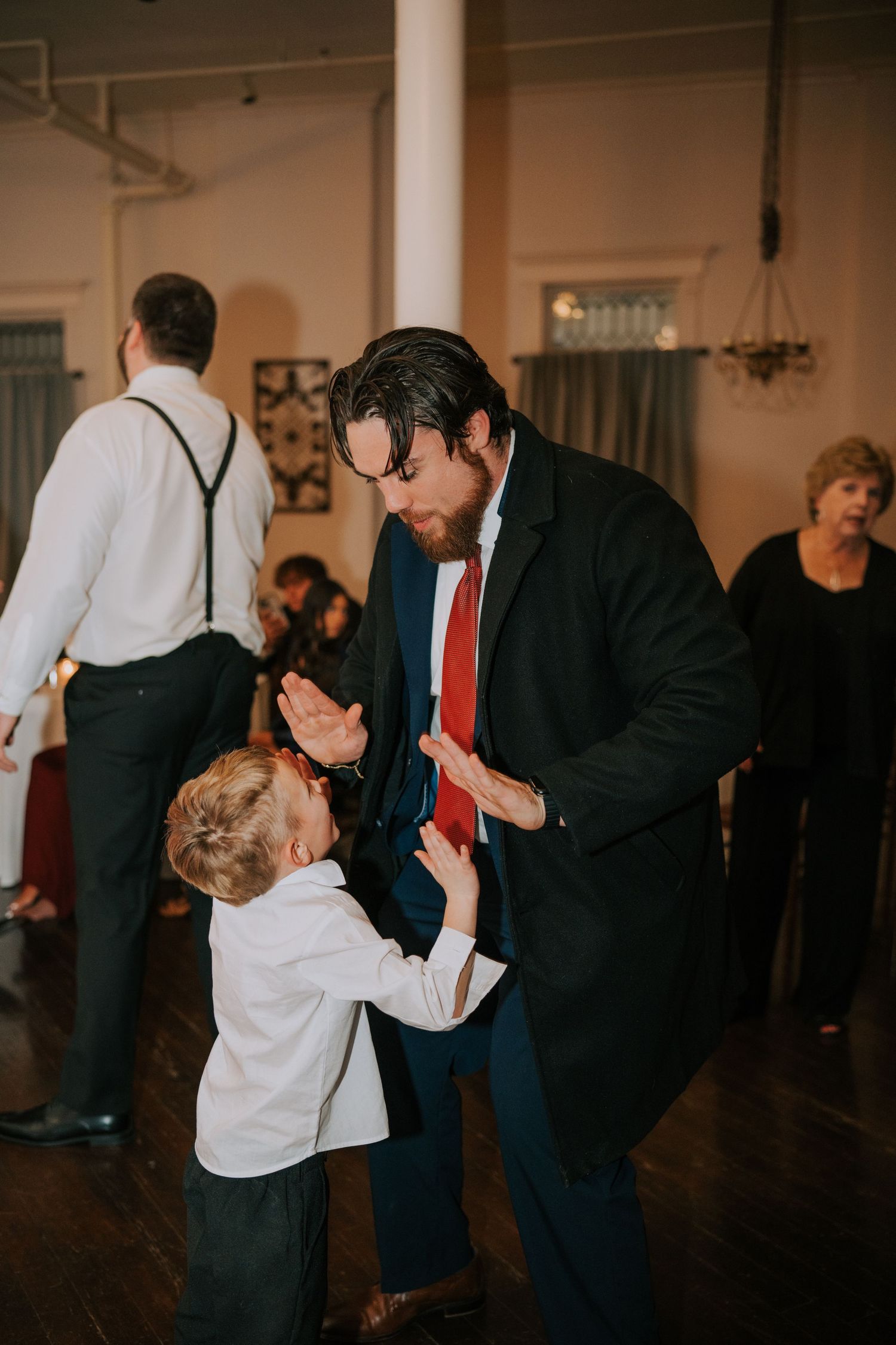 Wedding guest in suit dances with young child in suspenders on reception dance floor.
